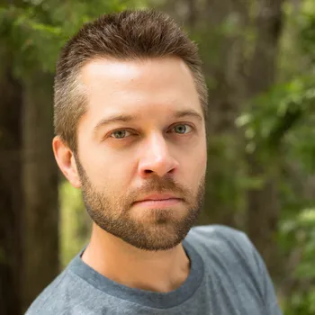 A young man with short brown hair wearing a blue t-shirt stands in a green, wooded outdoor area. 