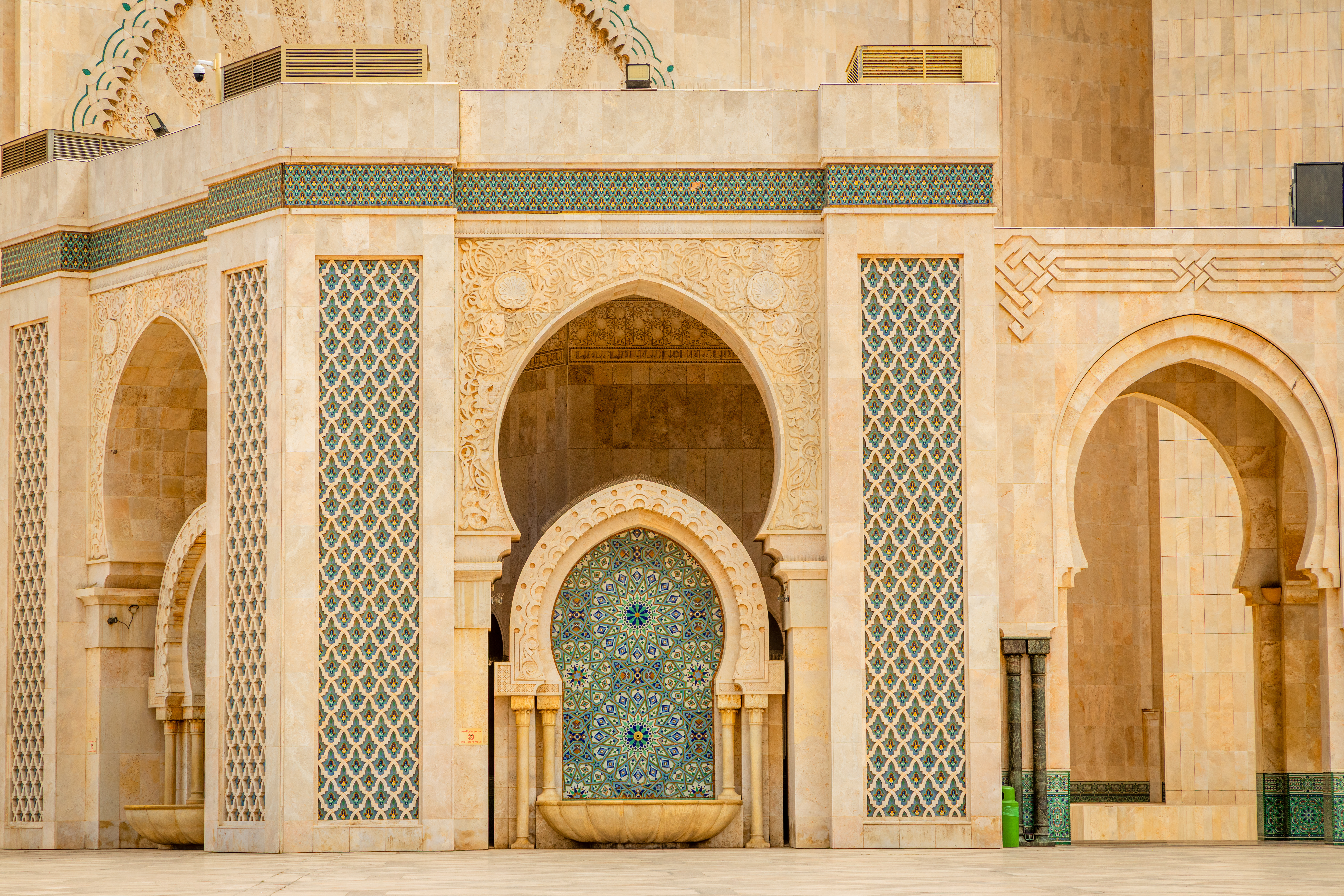 Pointed arches, carved stone, and a blue mosaic fountain panel create a symmetrical facade at Hassan II Mosque.