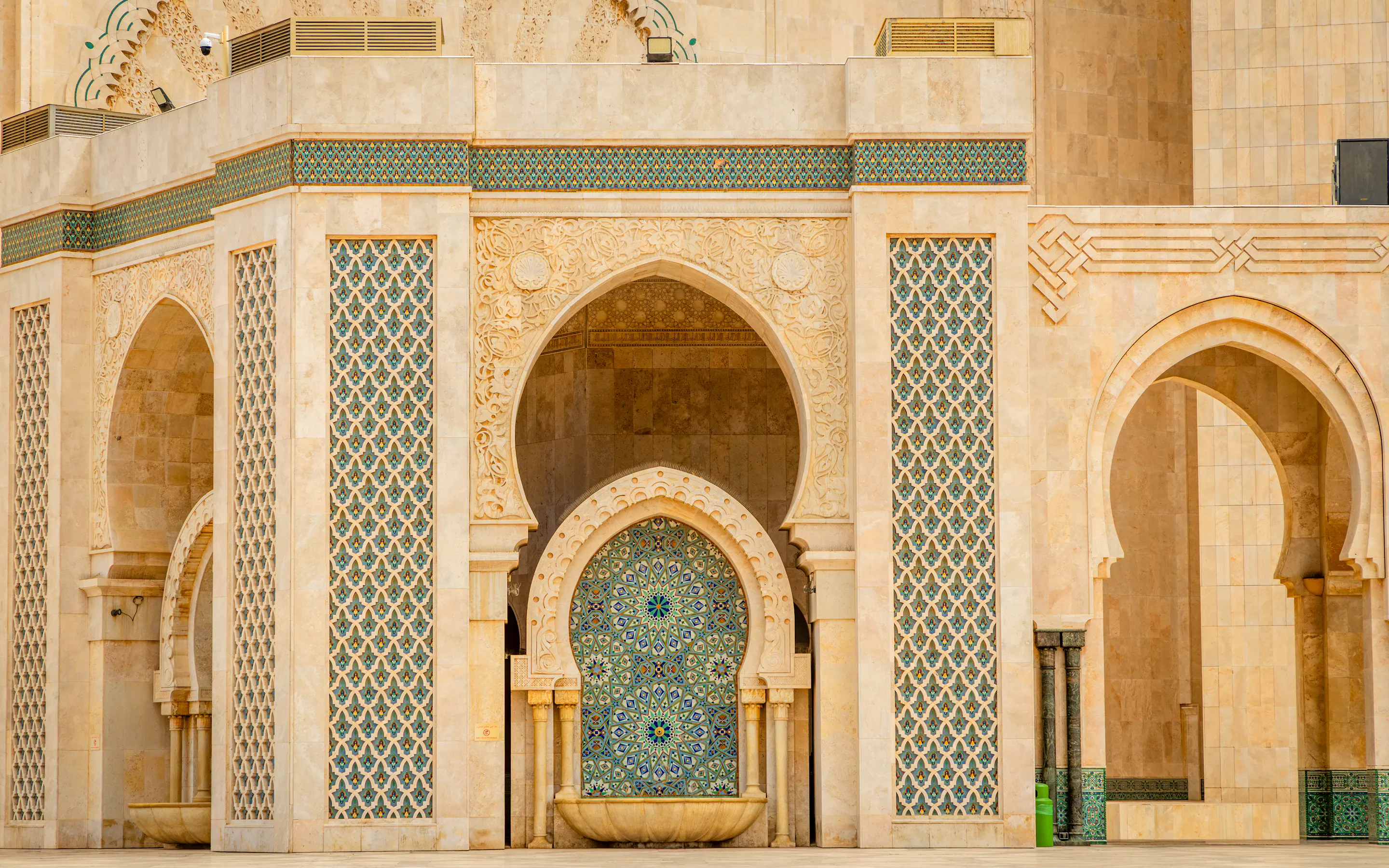 Pointed arches, carved stone, and a blue mosaic fountain panel create a symmetrical facade at Hassan II Mosque.