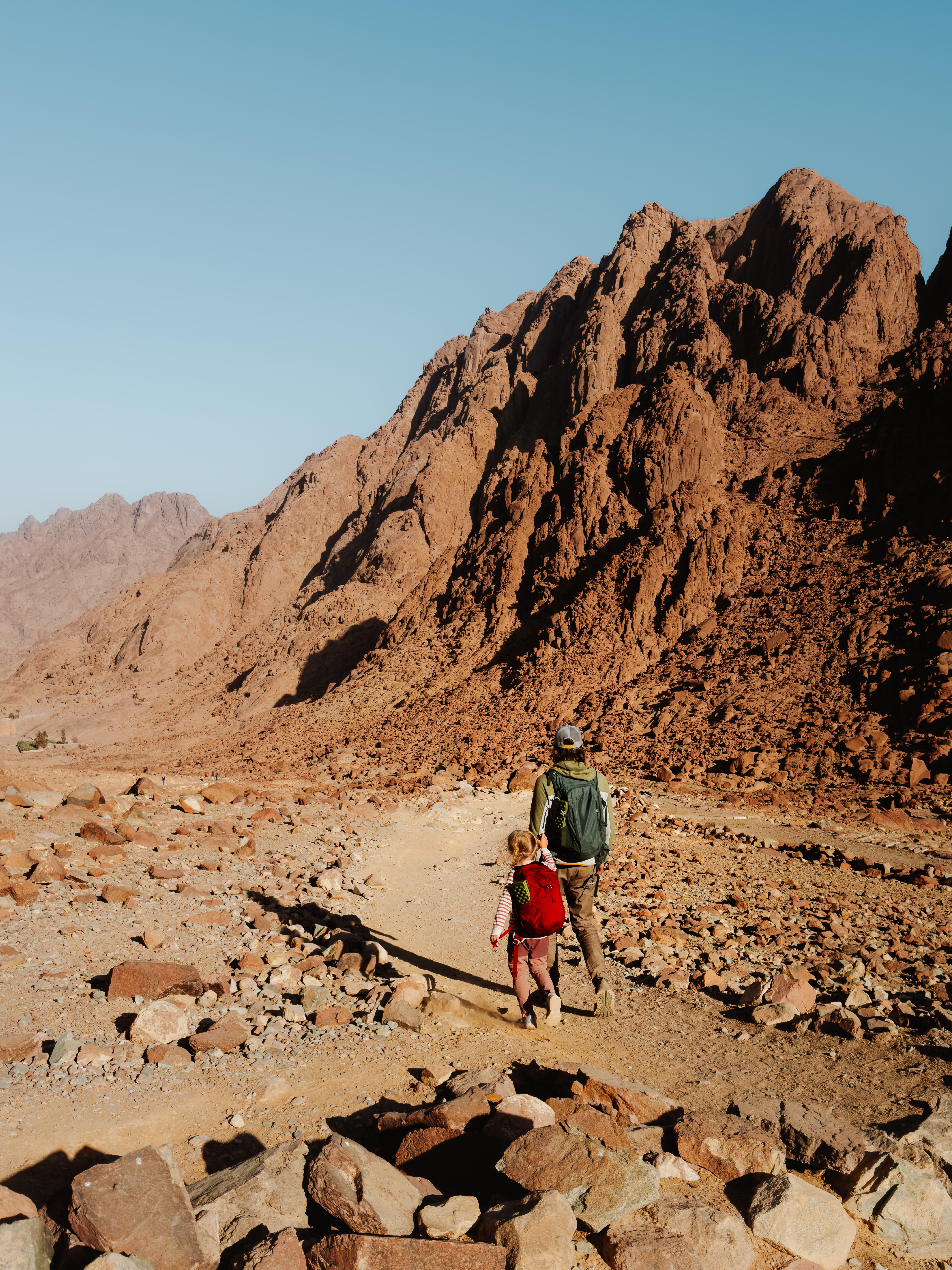 Two hikers walk a rocky trail through Sinai desert mountains, with steep red cliffs and a pale blue sky beyond.