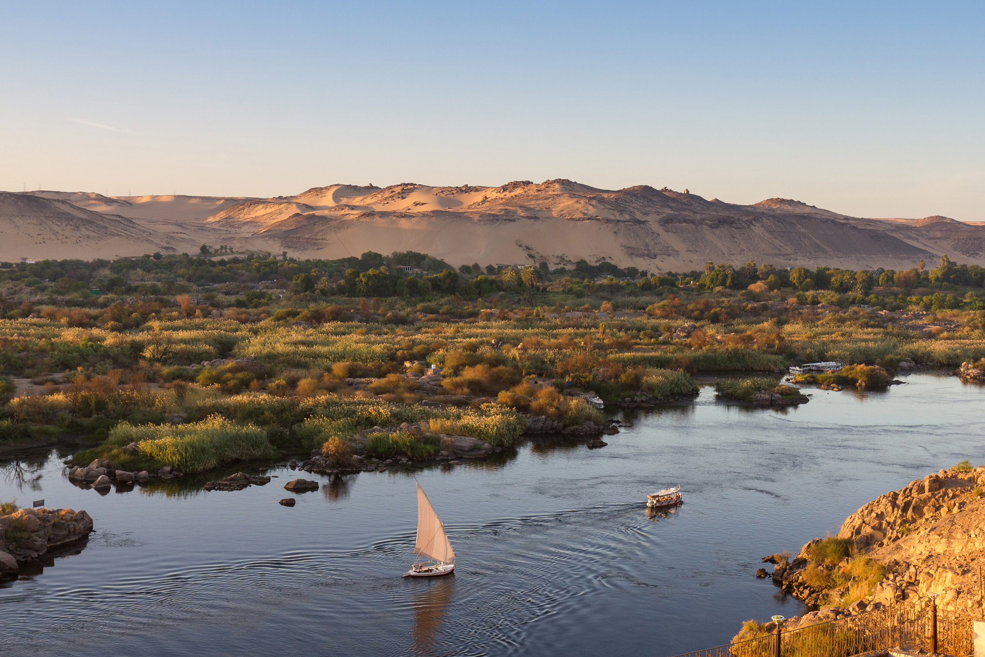A white sailboat moves along the Nile at sunset, with green riverbanks, calm water, and soft desert hills beyond.
