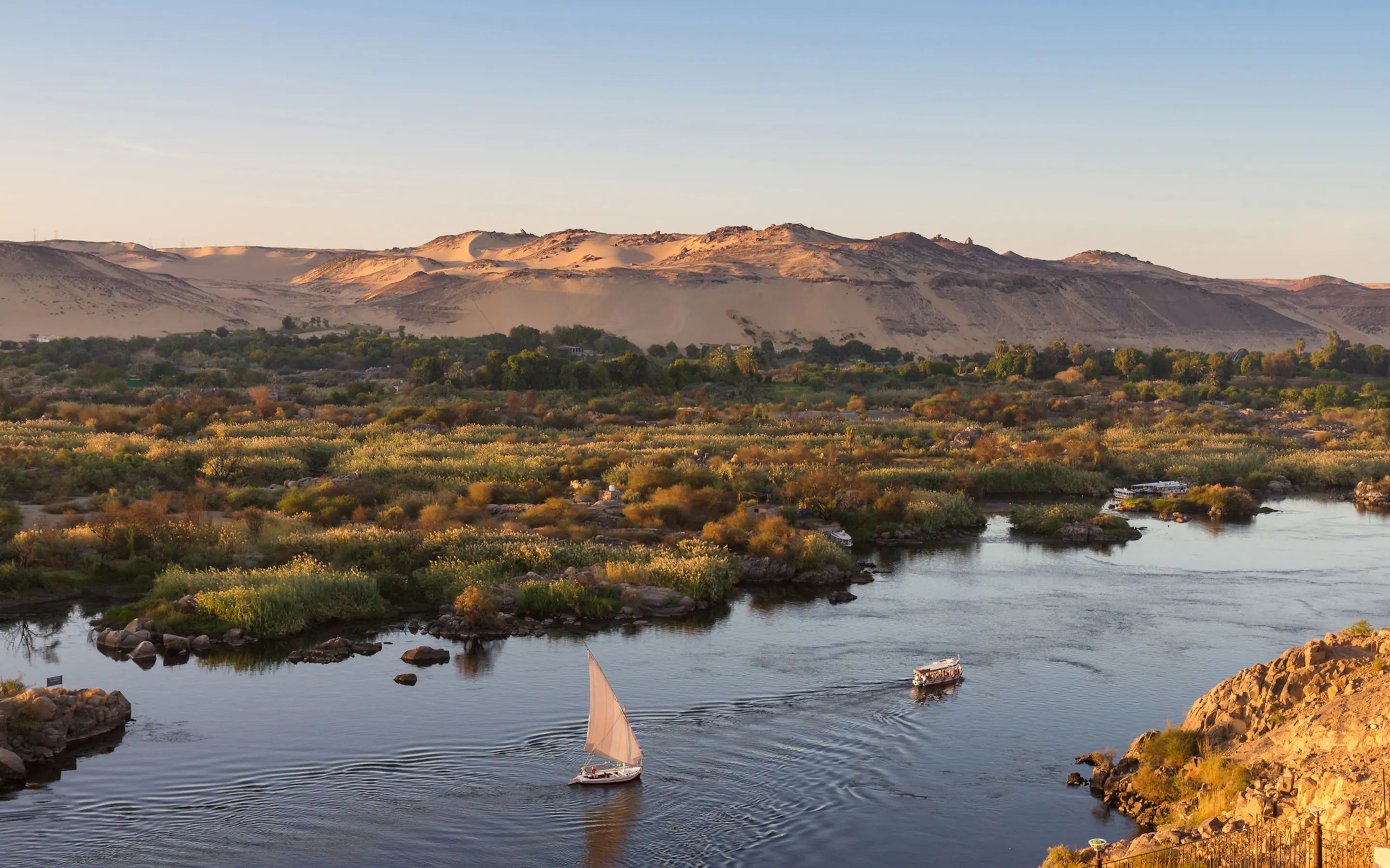 A white sailboat moves along the Nile at sunset, with green riverbanks, calm water, and soft desert hills beyond.