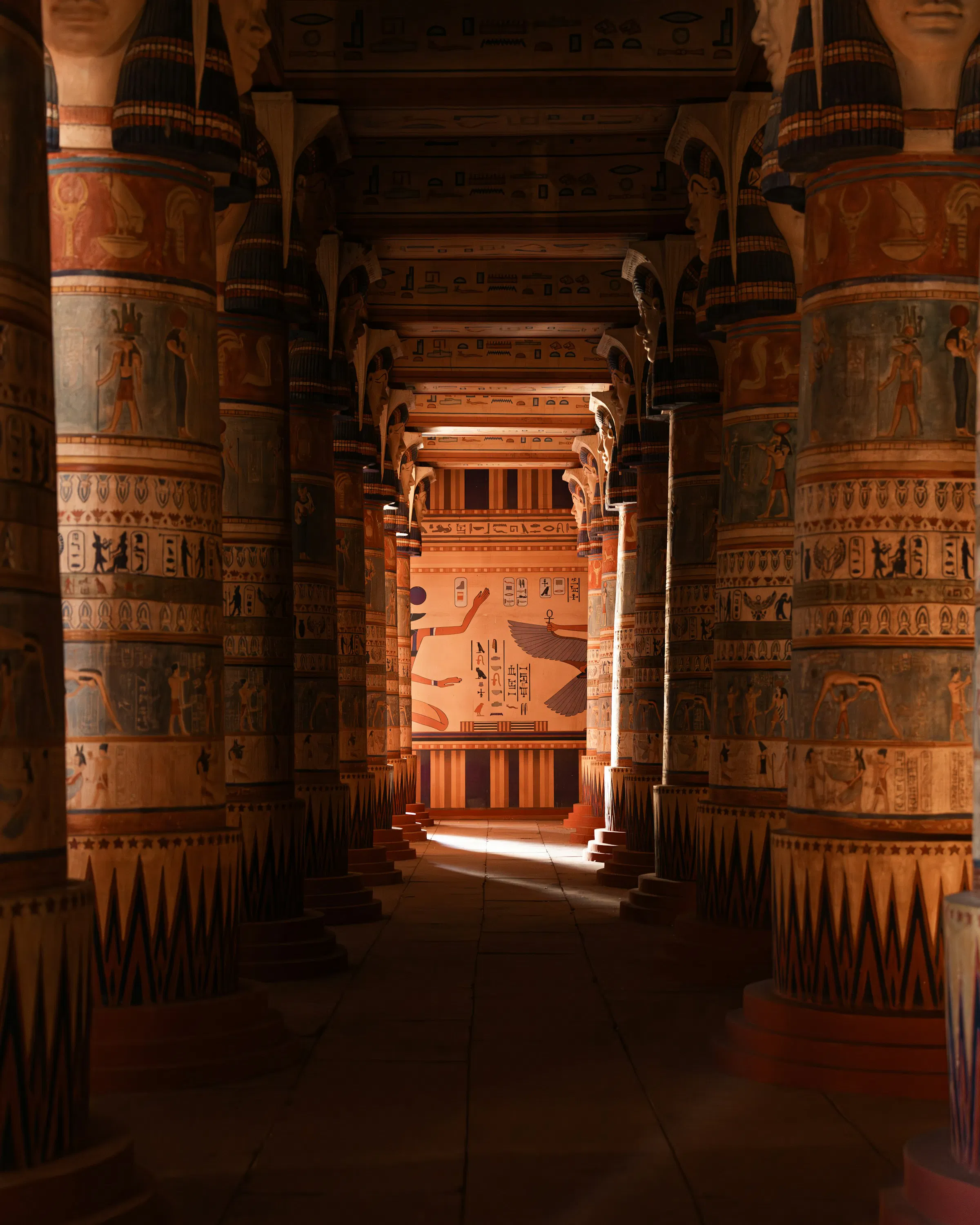 Painted columns line a dim temple hall, drawing the eye toward a bright doorway at the far end of the chamber.