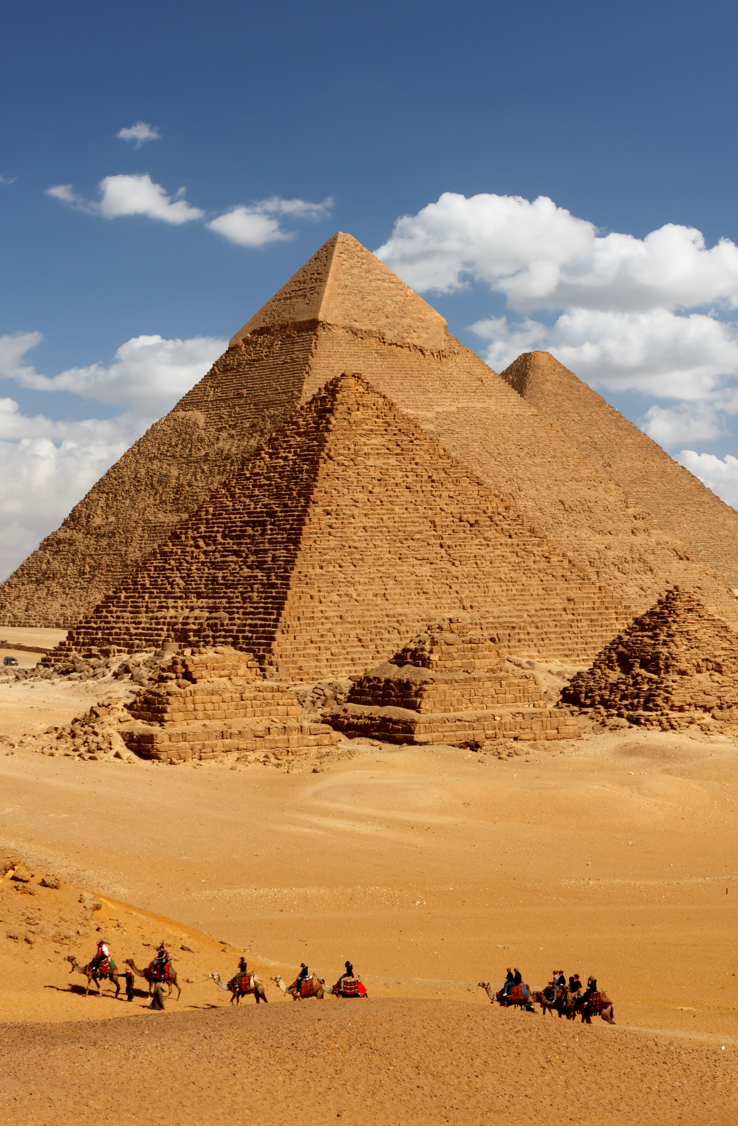 A line of camels crosses the sand before the Pyramids of Giza, with three sunlit monuments under scattered clouds.