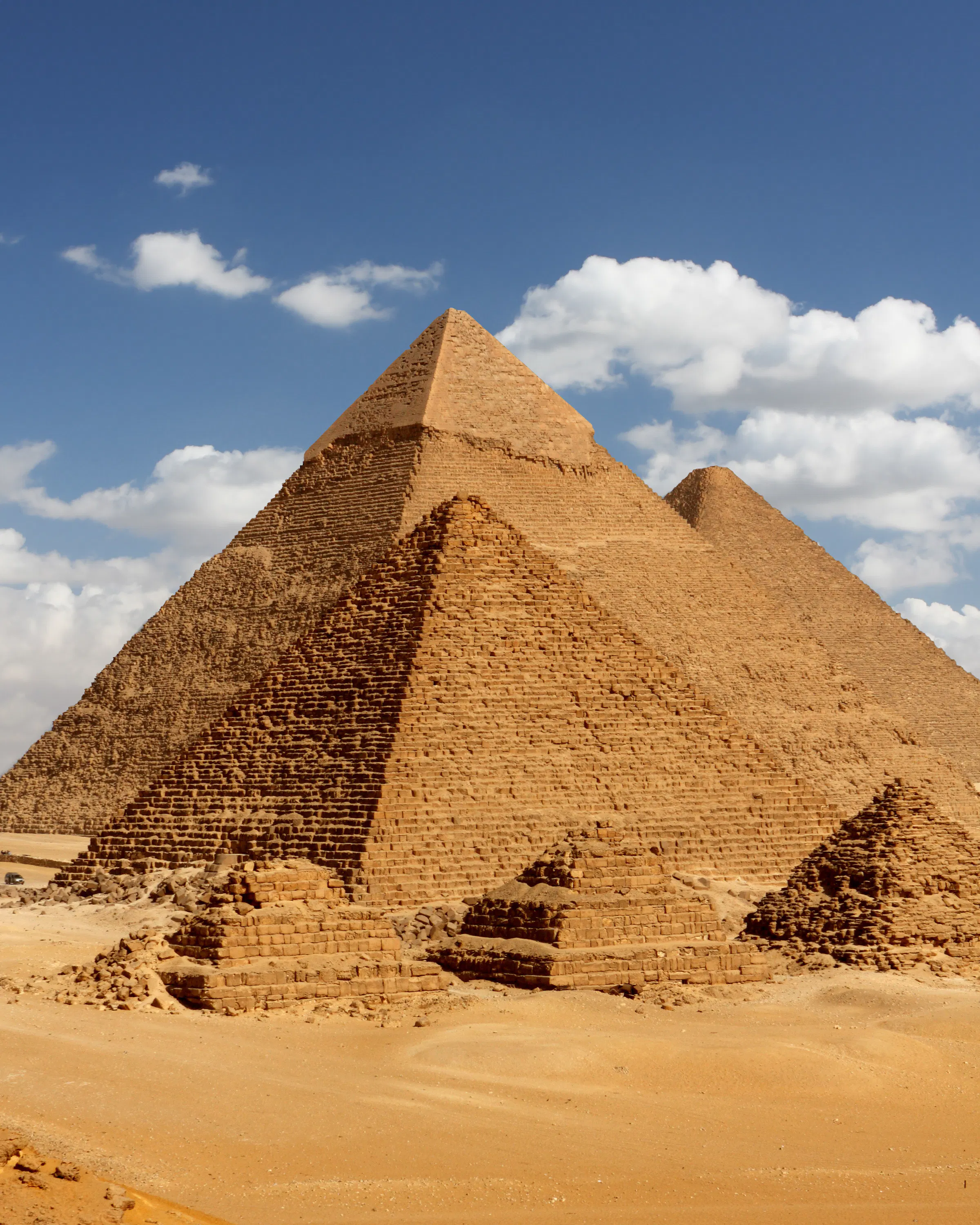 A line of camels crosses the sand before the Pyramids of Giza, with three sunlit monuments under scattered clouds.
