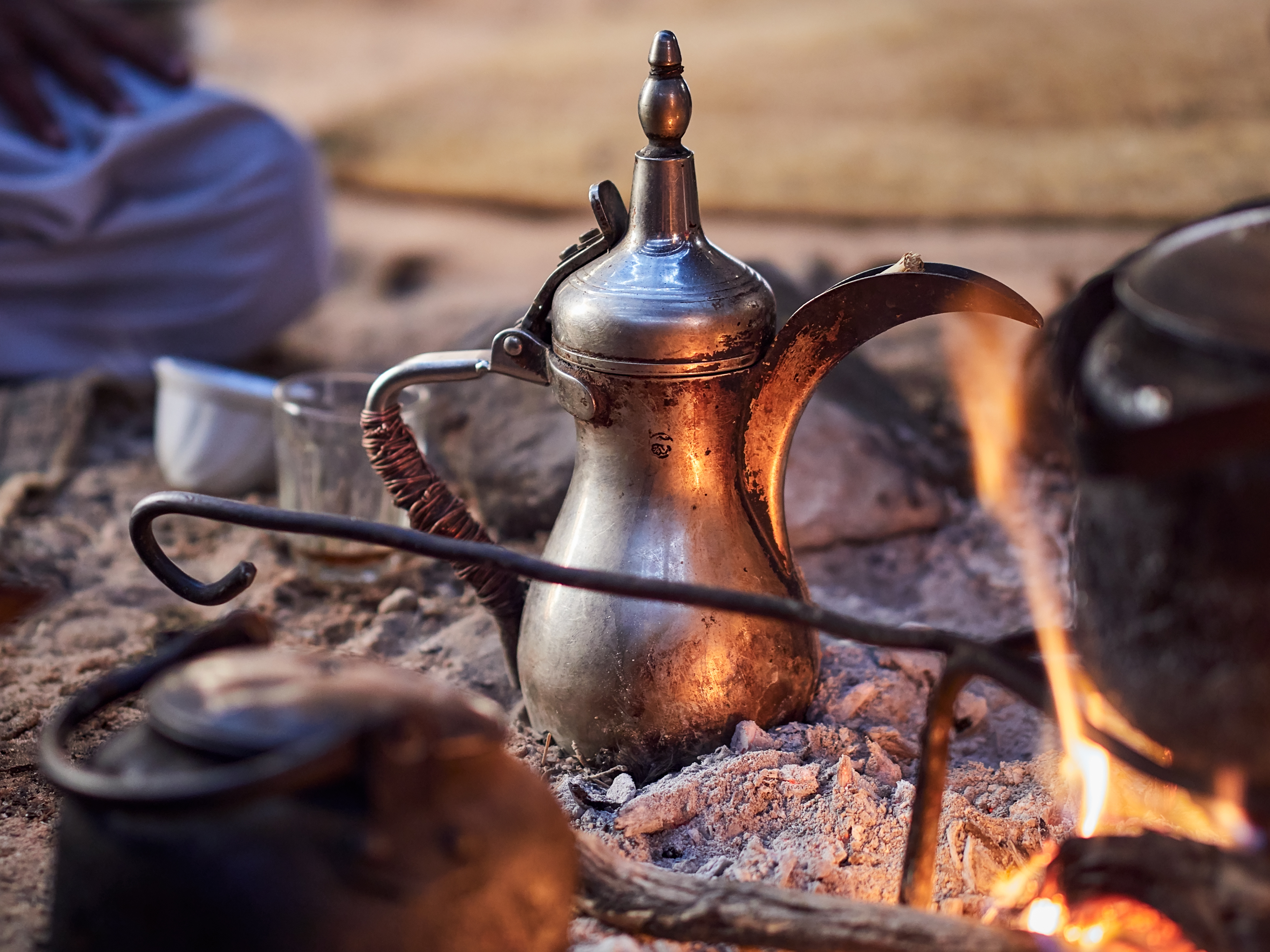 A metal coffee pot sits in warm embers beside open flames, with cups and kettles gathered close in the sand.