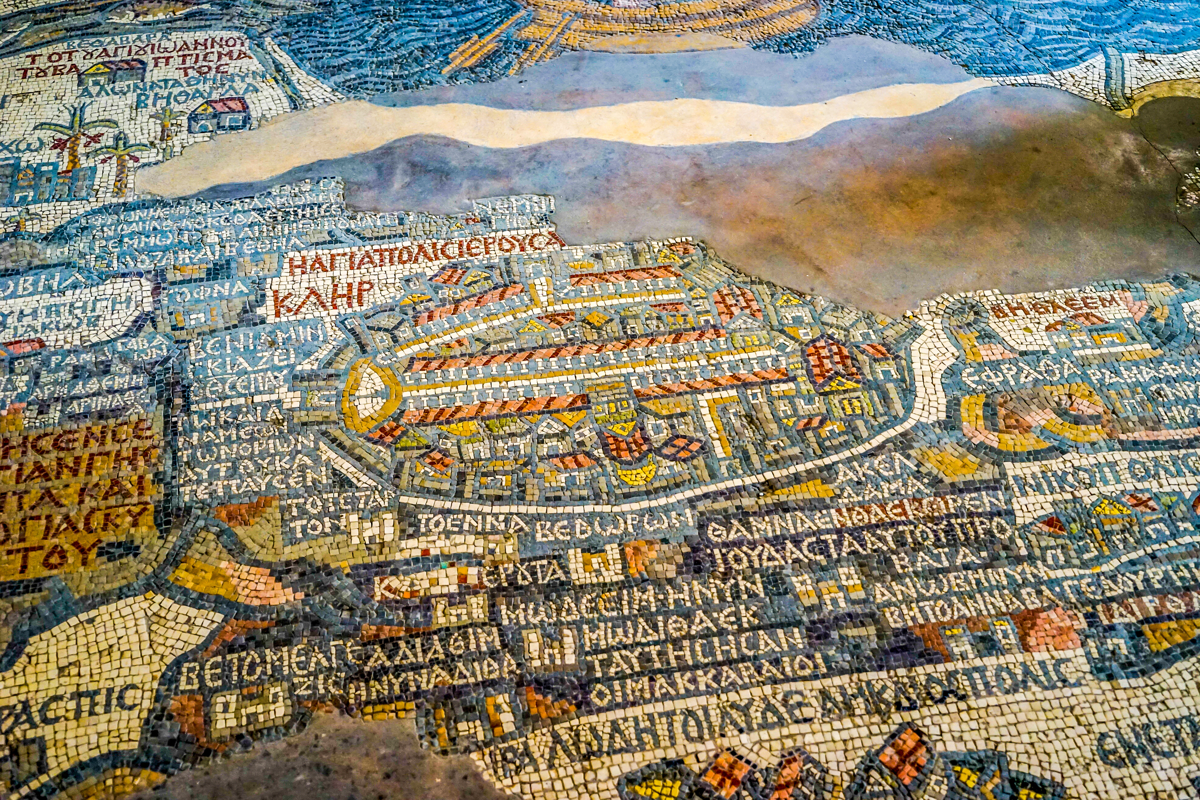 Colored stone tesserae form the ancient Madaba mosaic map, with Greek script and stylized streets in view.