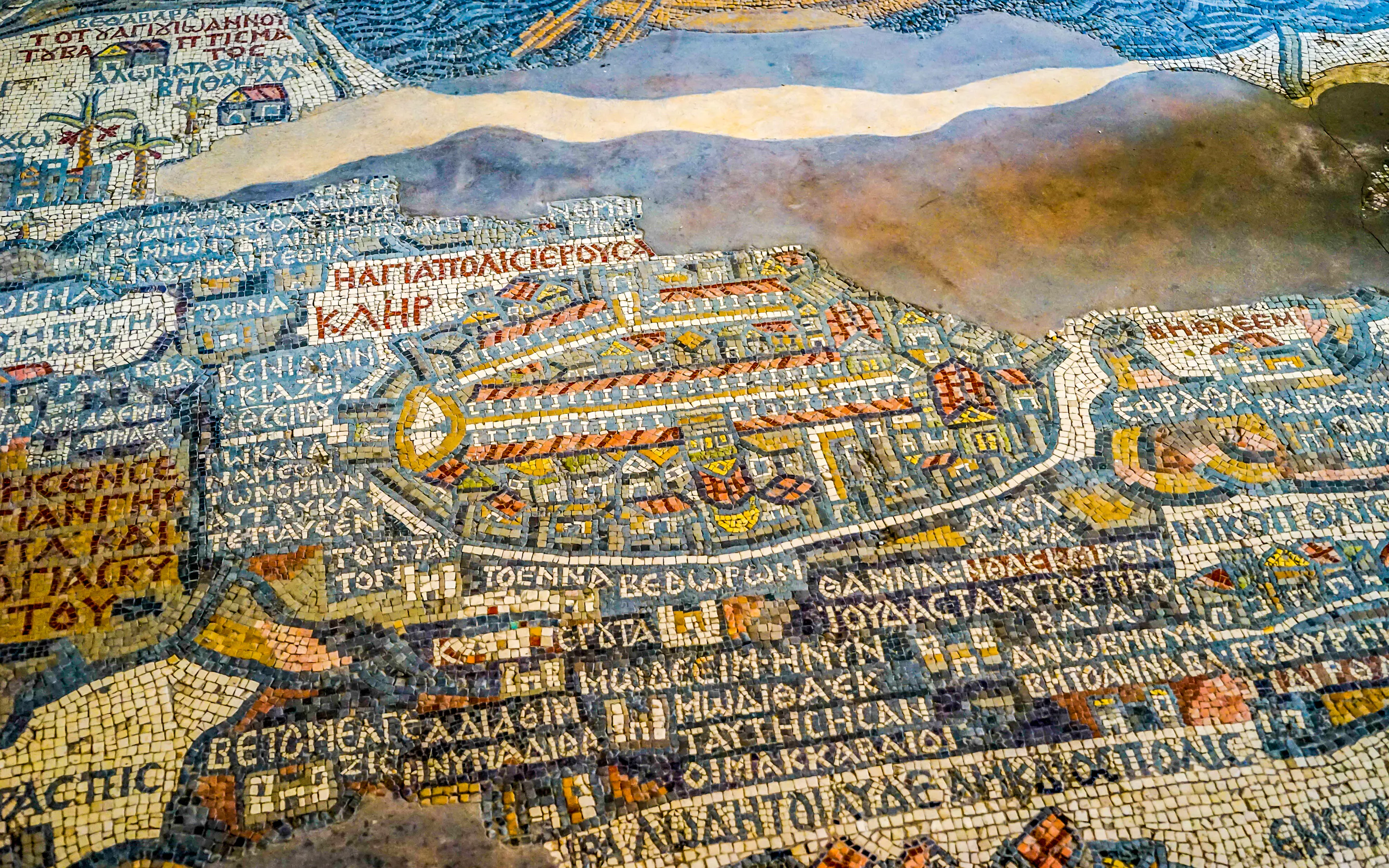 Colored stone tesserae form the ancient Madaba mosaic map, with Greek script and stylized streets in view.