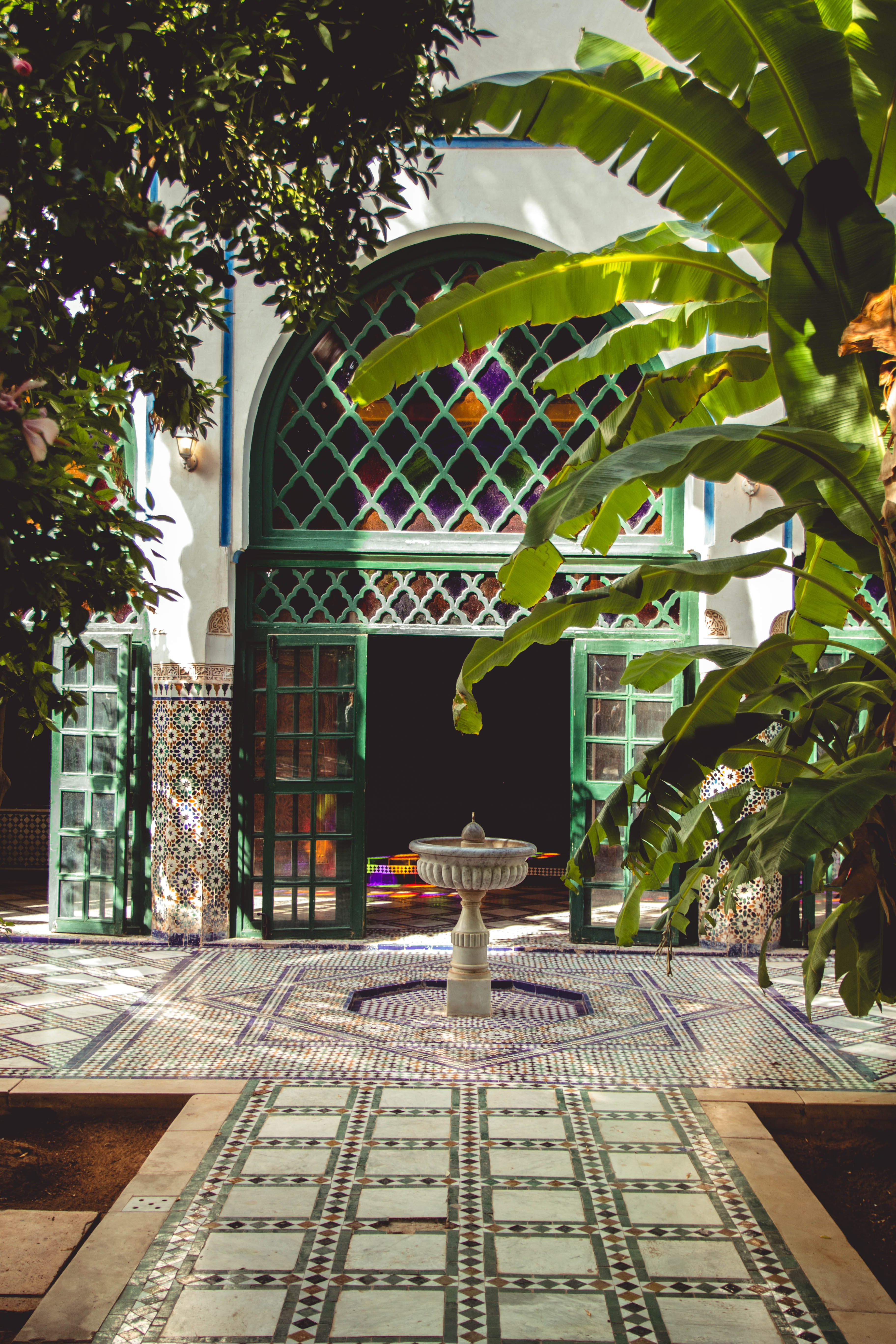 A tiled courtyard frames a stone fountain beneath arched windows, with broad green leaves casting patterned shade.