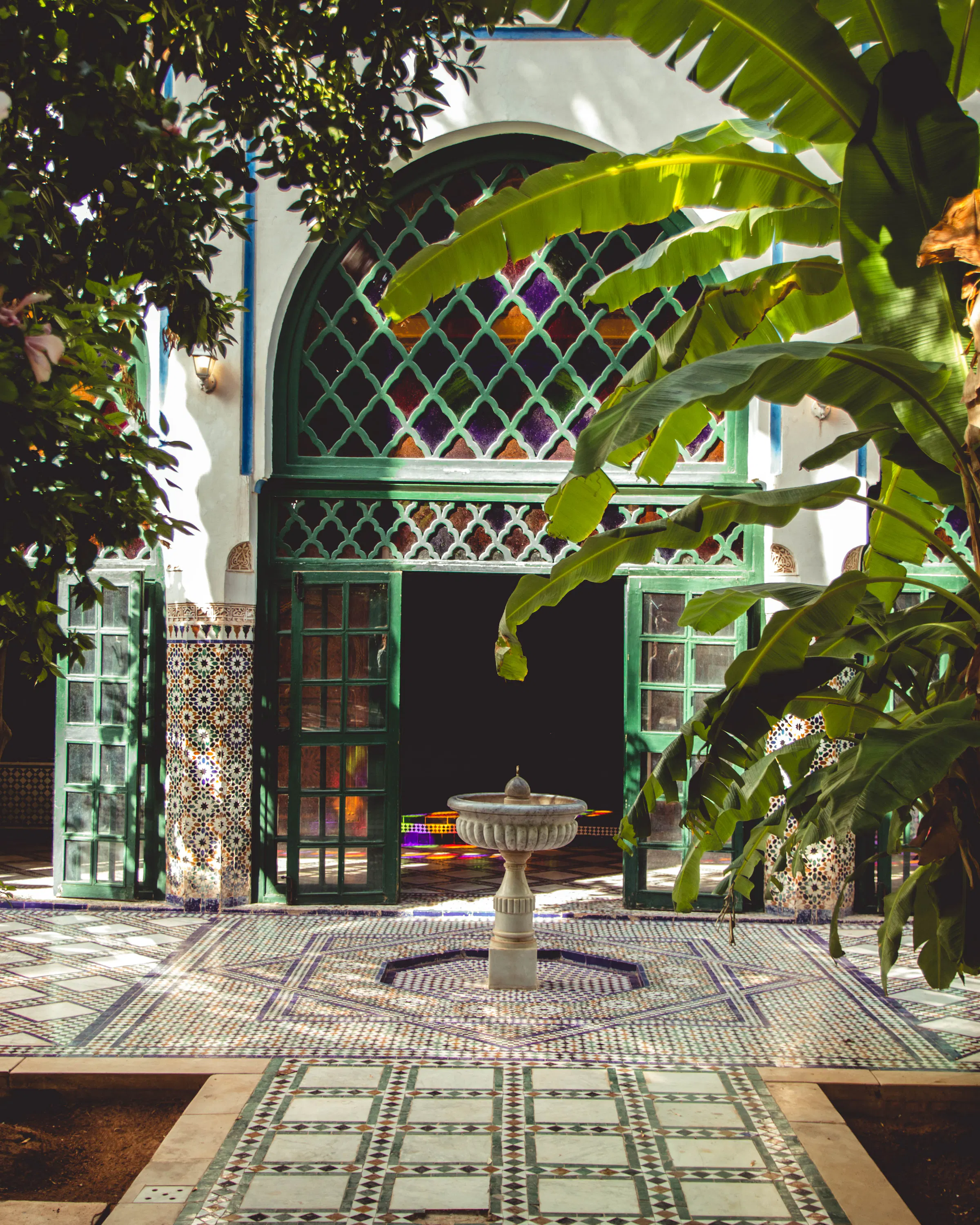 A tiled courtyard frames a stone fountain beneath arched windows, with broad green leaves casting patterned shade.