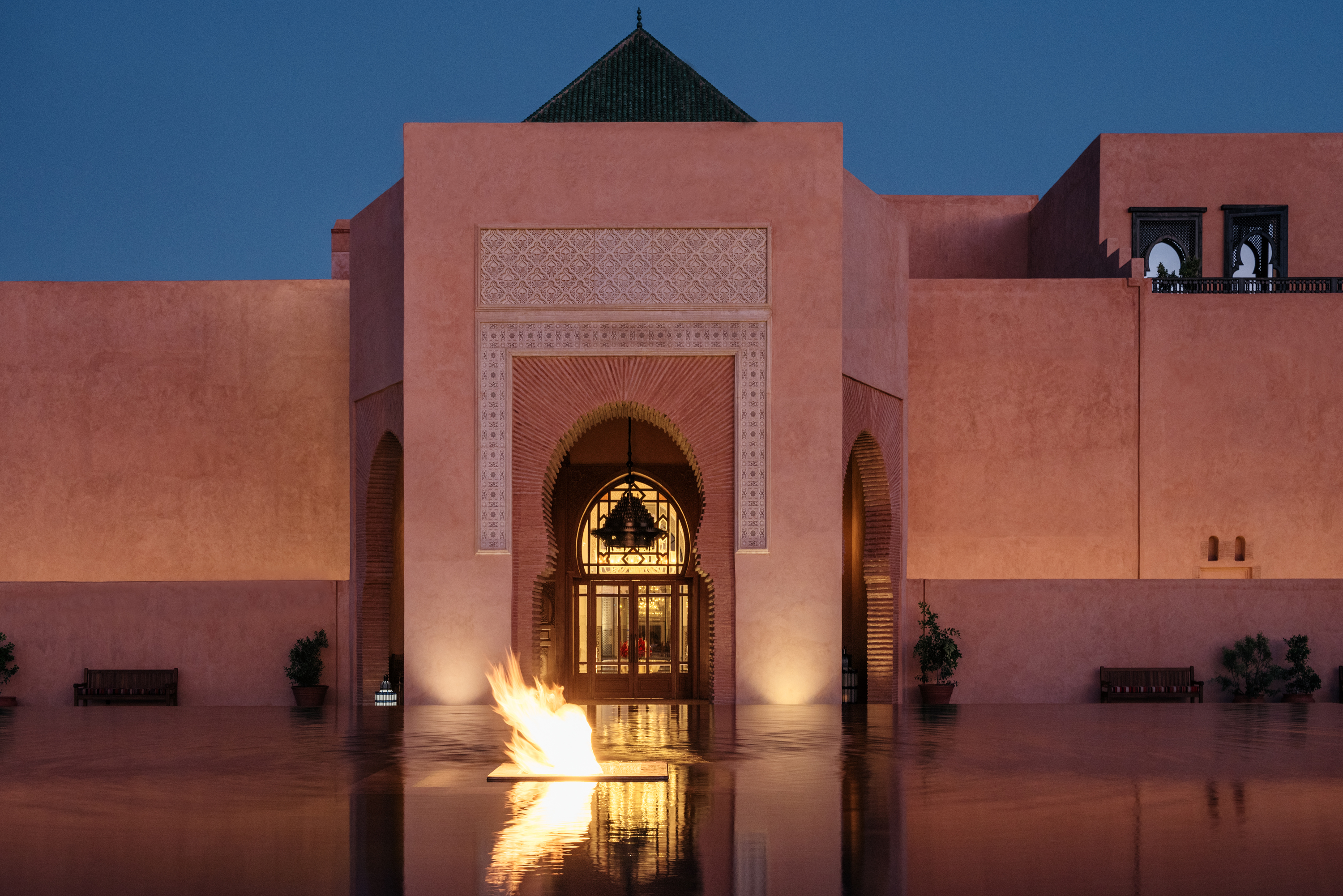 The illuminated entrance of The Oberoi, Marrakech rises behind a reflective pool and fire feature at dusk.