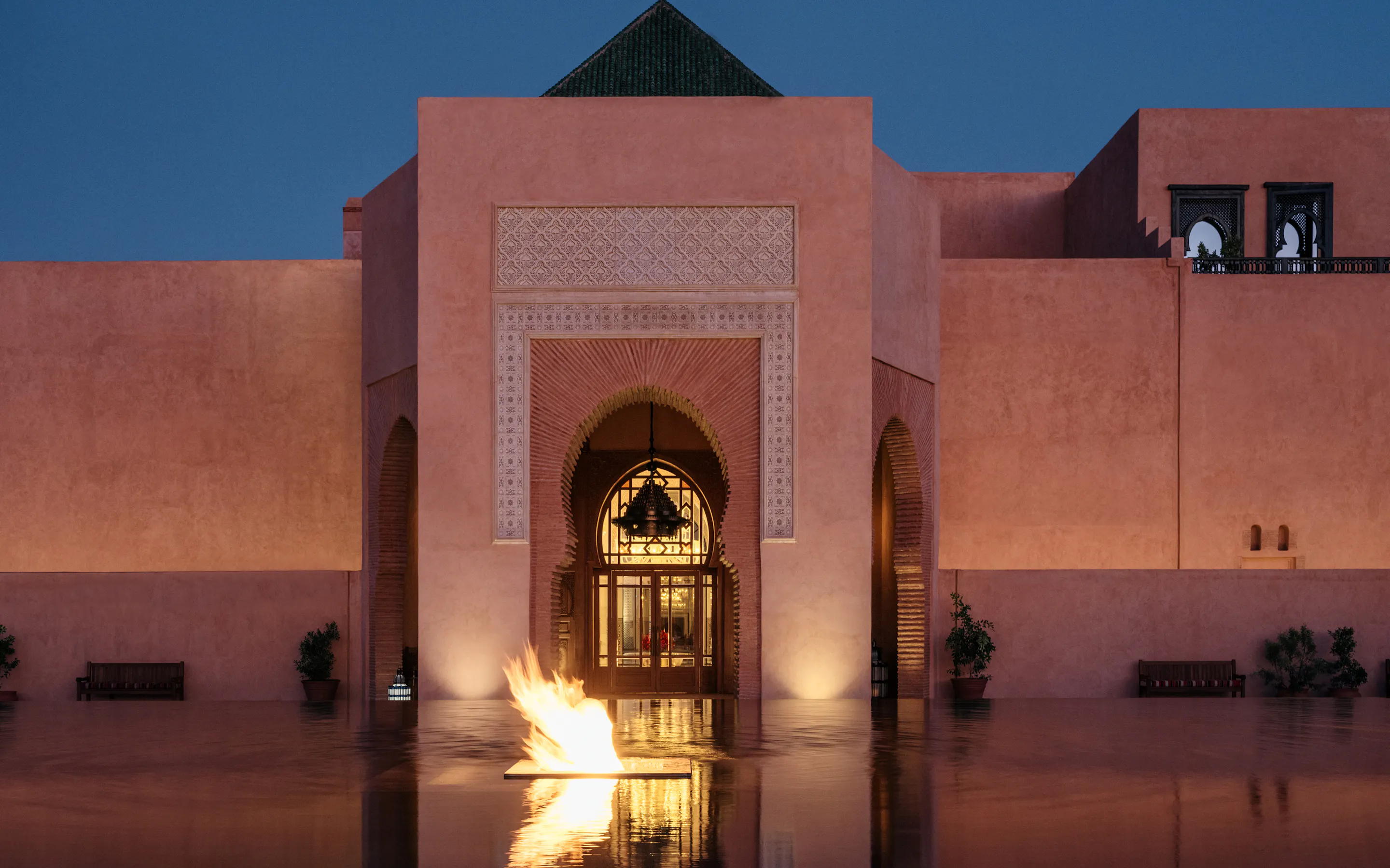 The illuminated entrance of The Oberoi, Marrakech rises behind a reflective pool and fire feature at dusk.