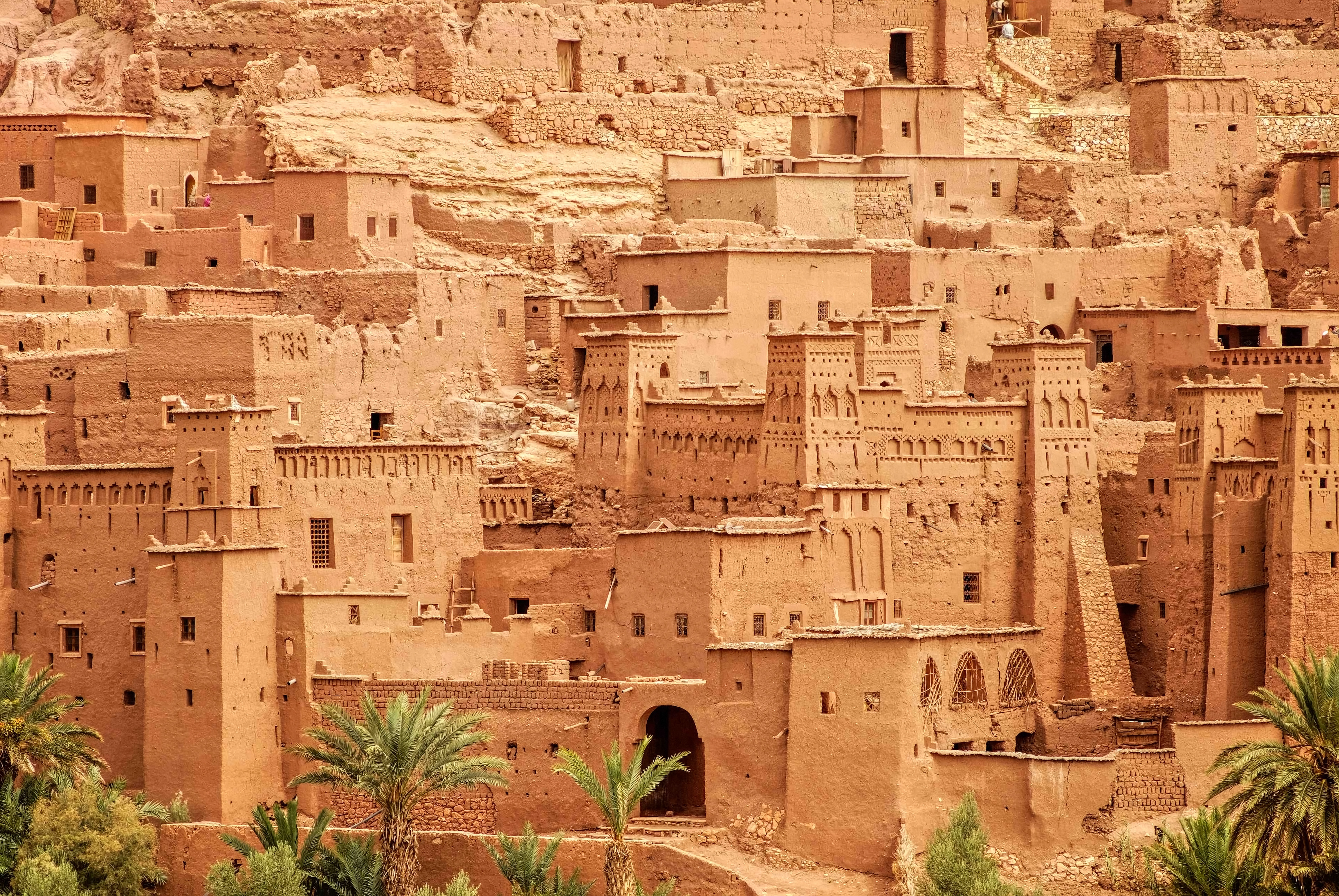 Layered clay towers and narrow windows rise above palms at Ait Benhaddou, revealing the ksar's dense earthen texture.