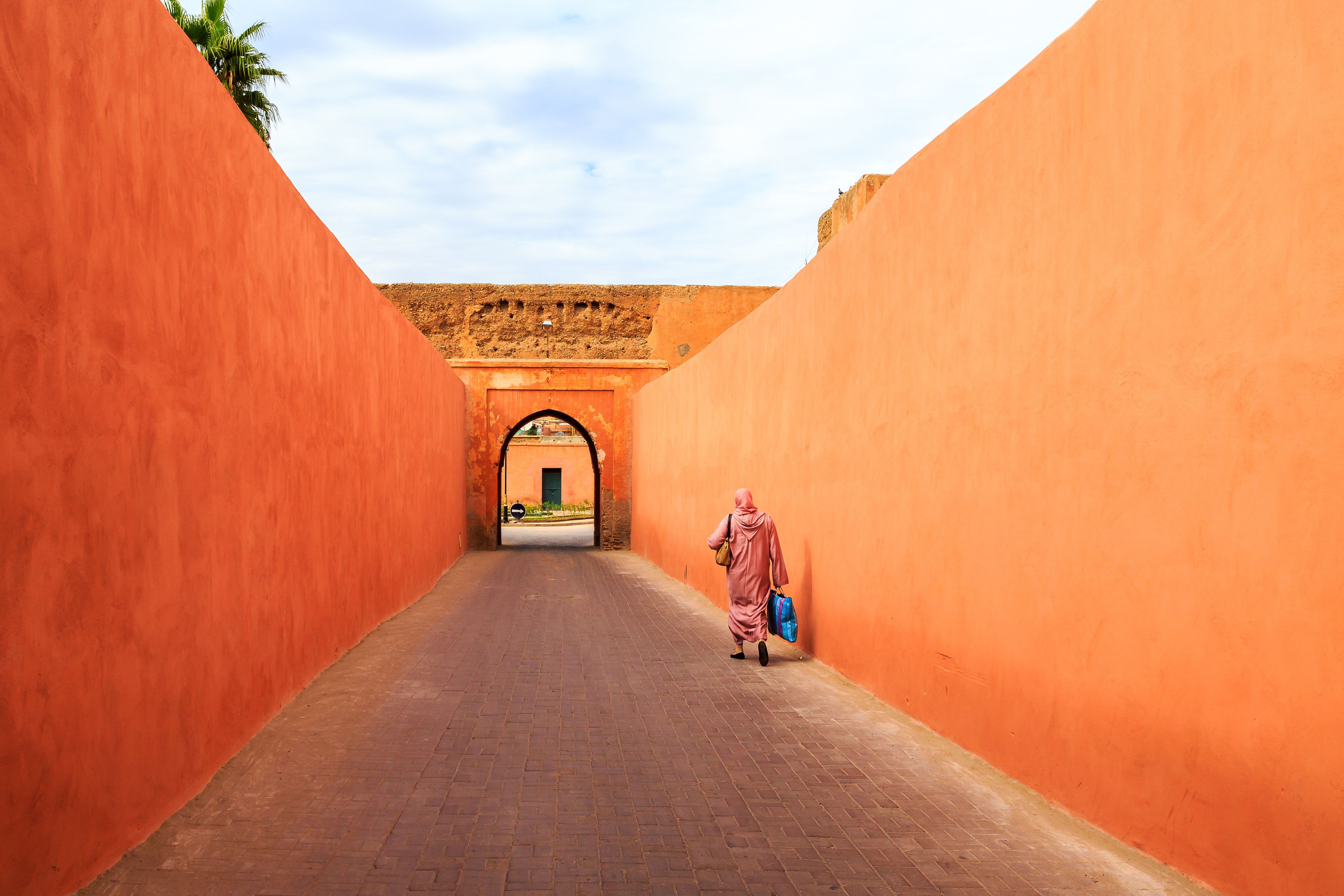 A woman walks along a terracotta-walled alley in Marrakech, where a pointed arch frames the sunlit lane ahead.