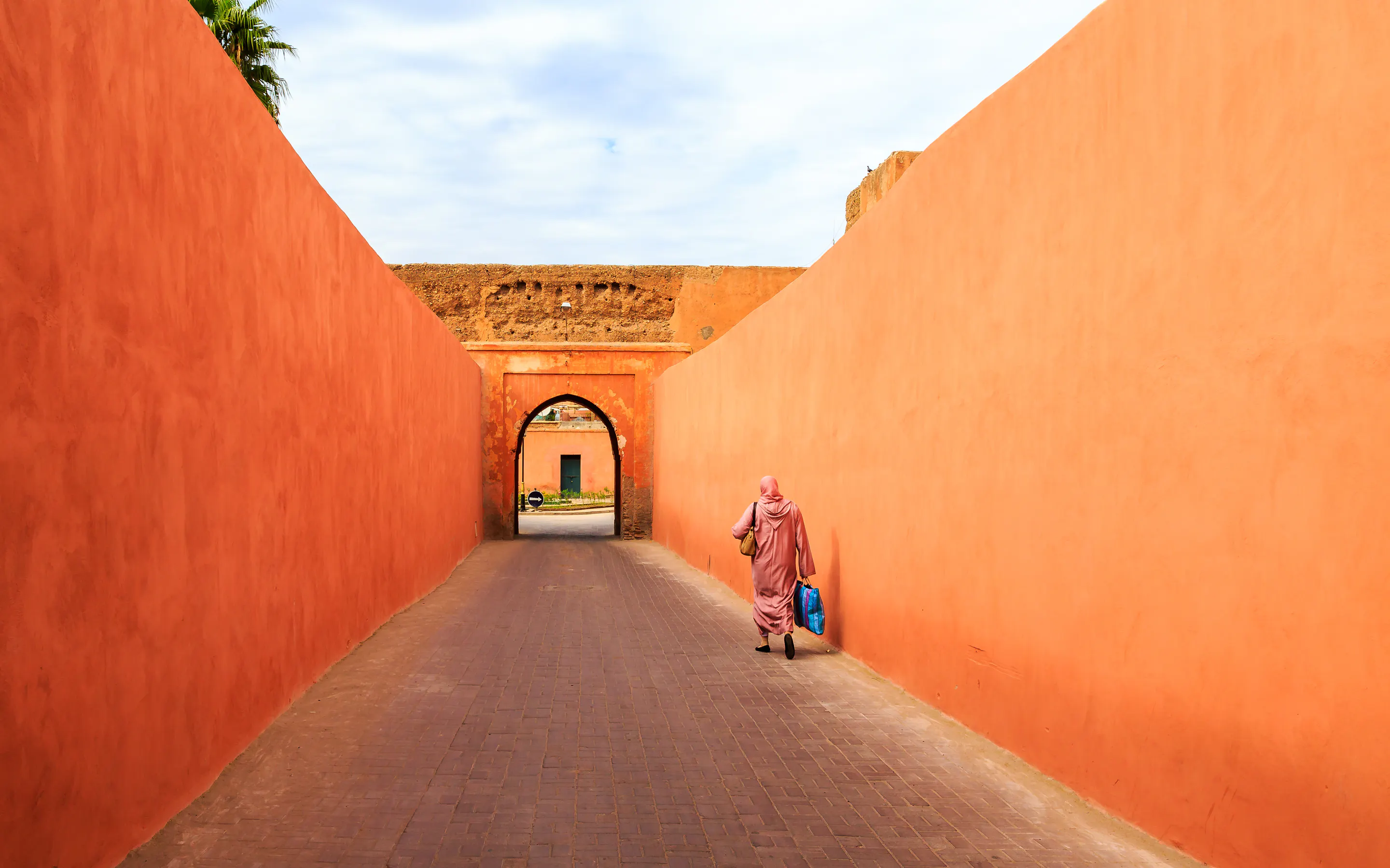 A woman walks along a terracotta-walled alley in Marrakech, where a pointed arch frames the sunlit lane ahead.
