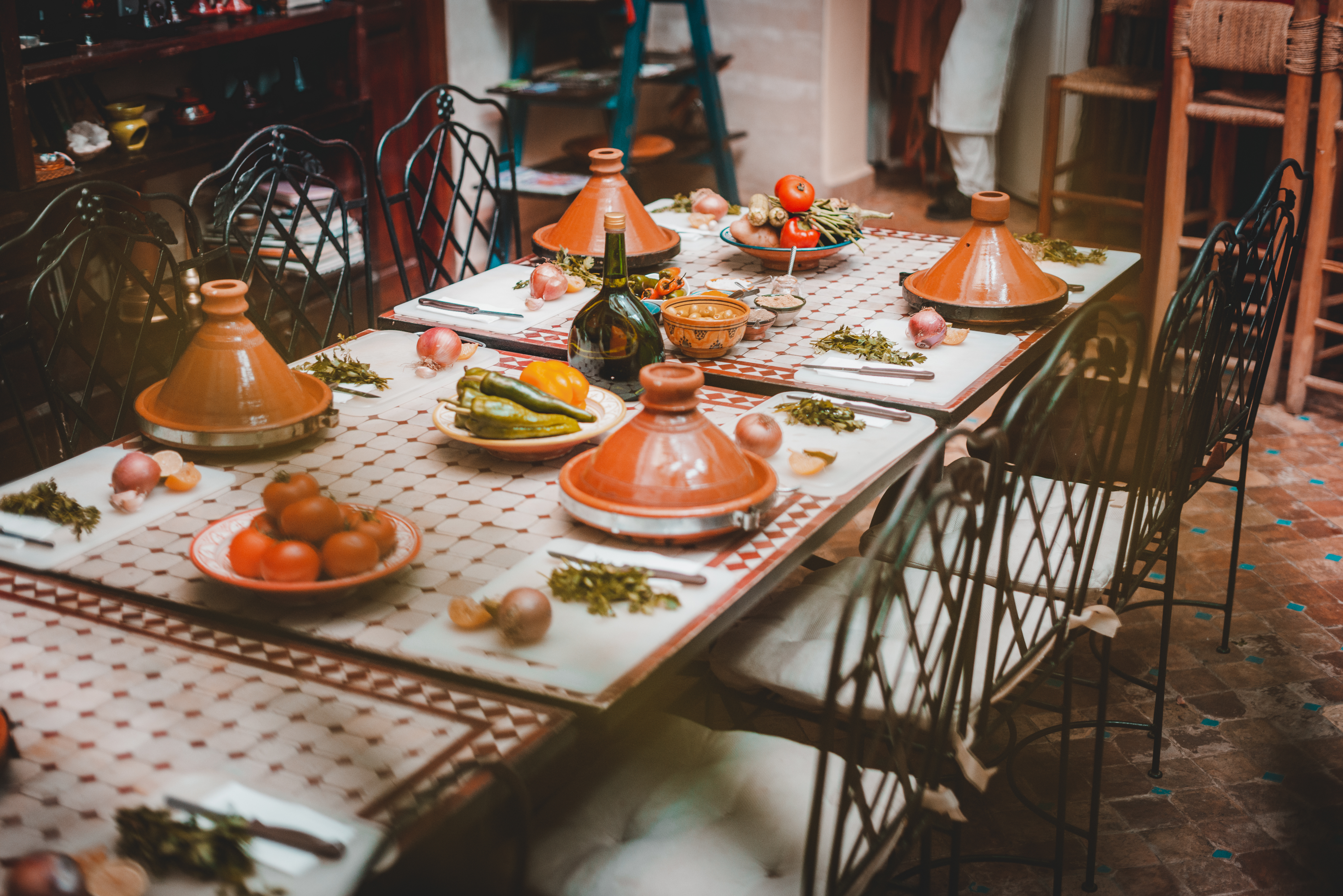 Tagine pots and fresh vegetables line a tiled table set for a Moroccan meal, with warm light softening the room.