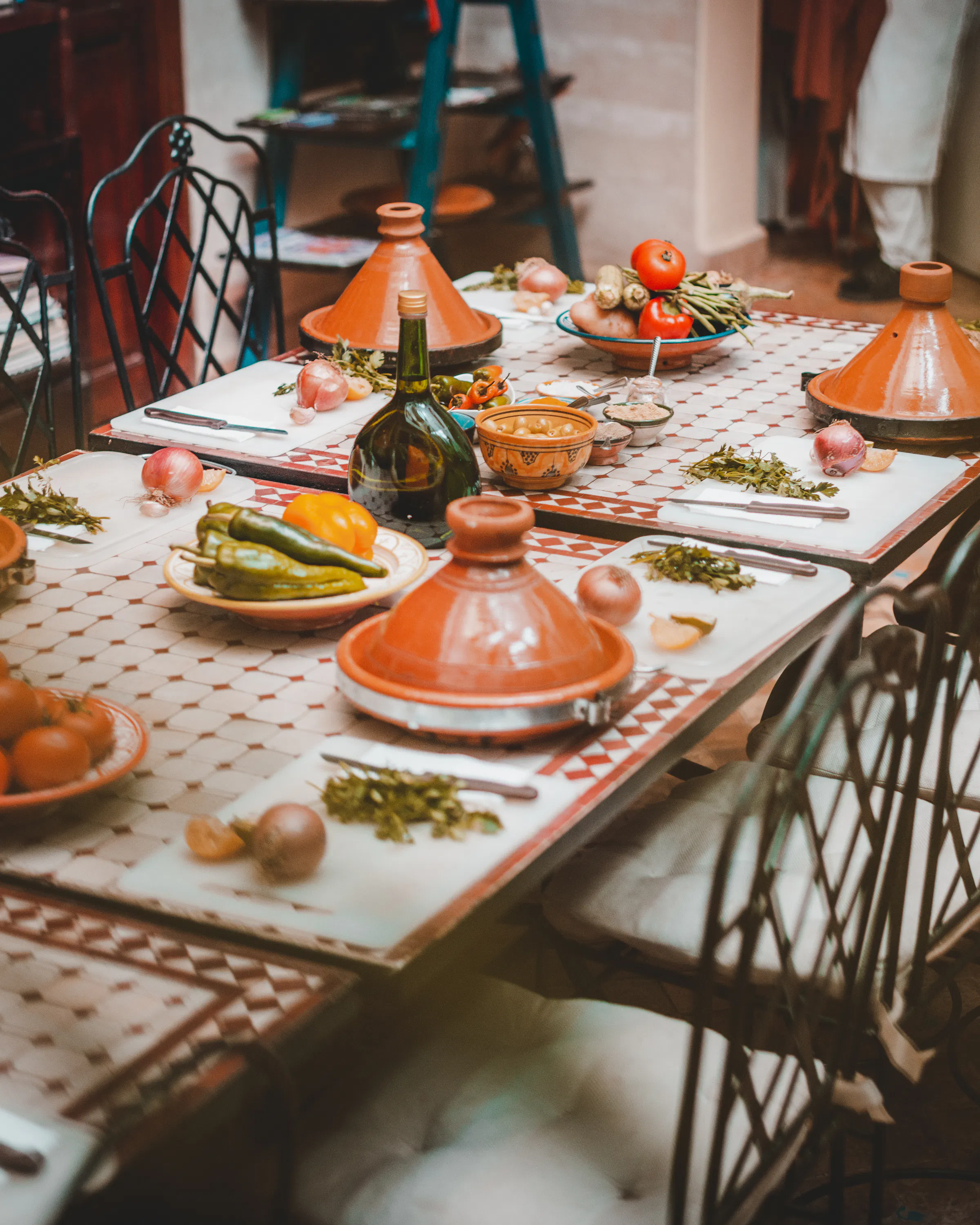 Tagine pots and fresh vegetables line a tiled table set for a Moroccan meal, with warm light softening the room.