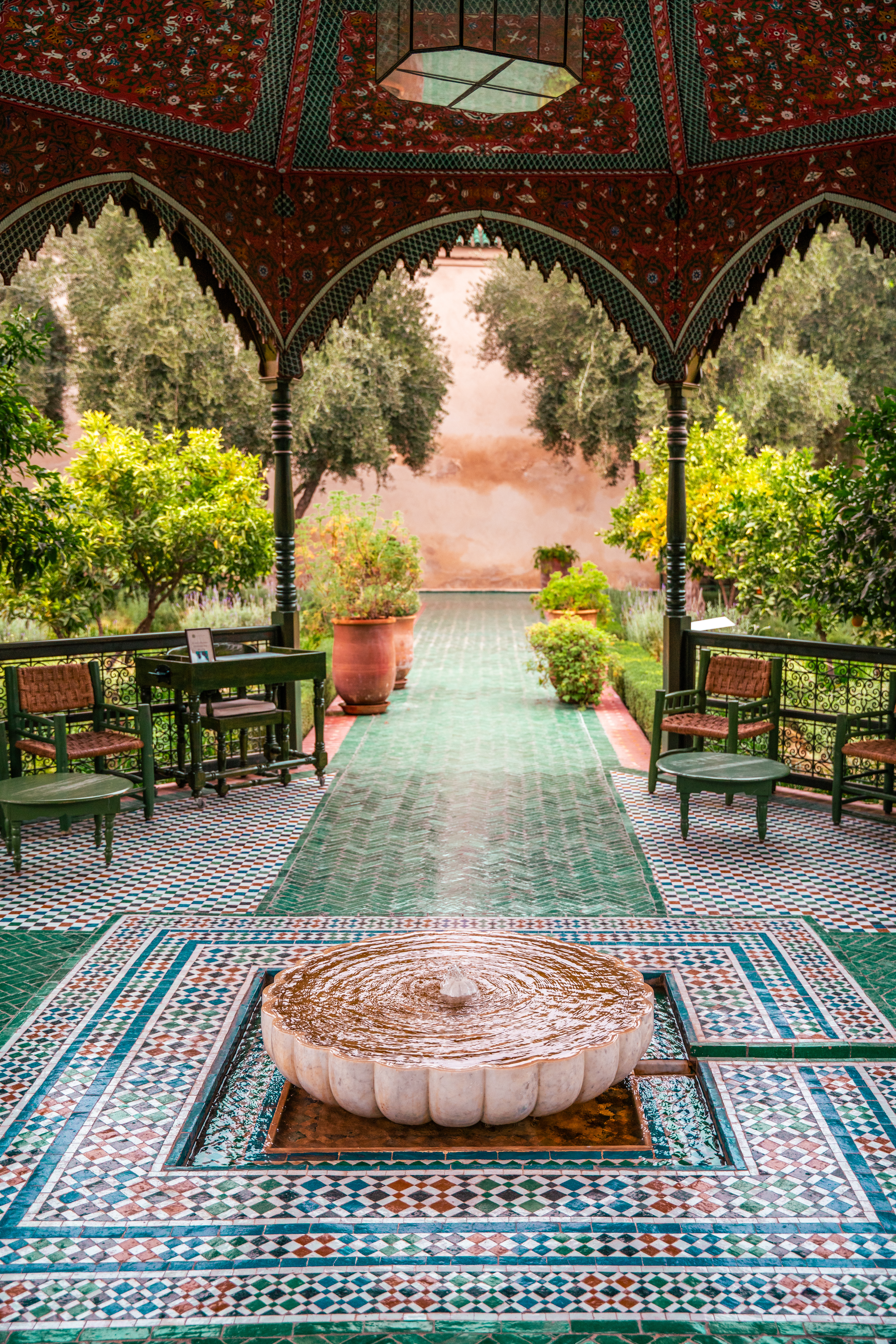 A tiled fountain ripples beneath a patterned pavilion in Marrakech, bordered by green chairs and a formal garden.