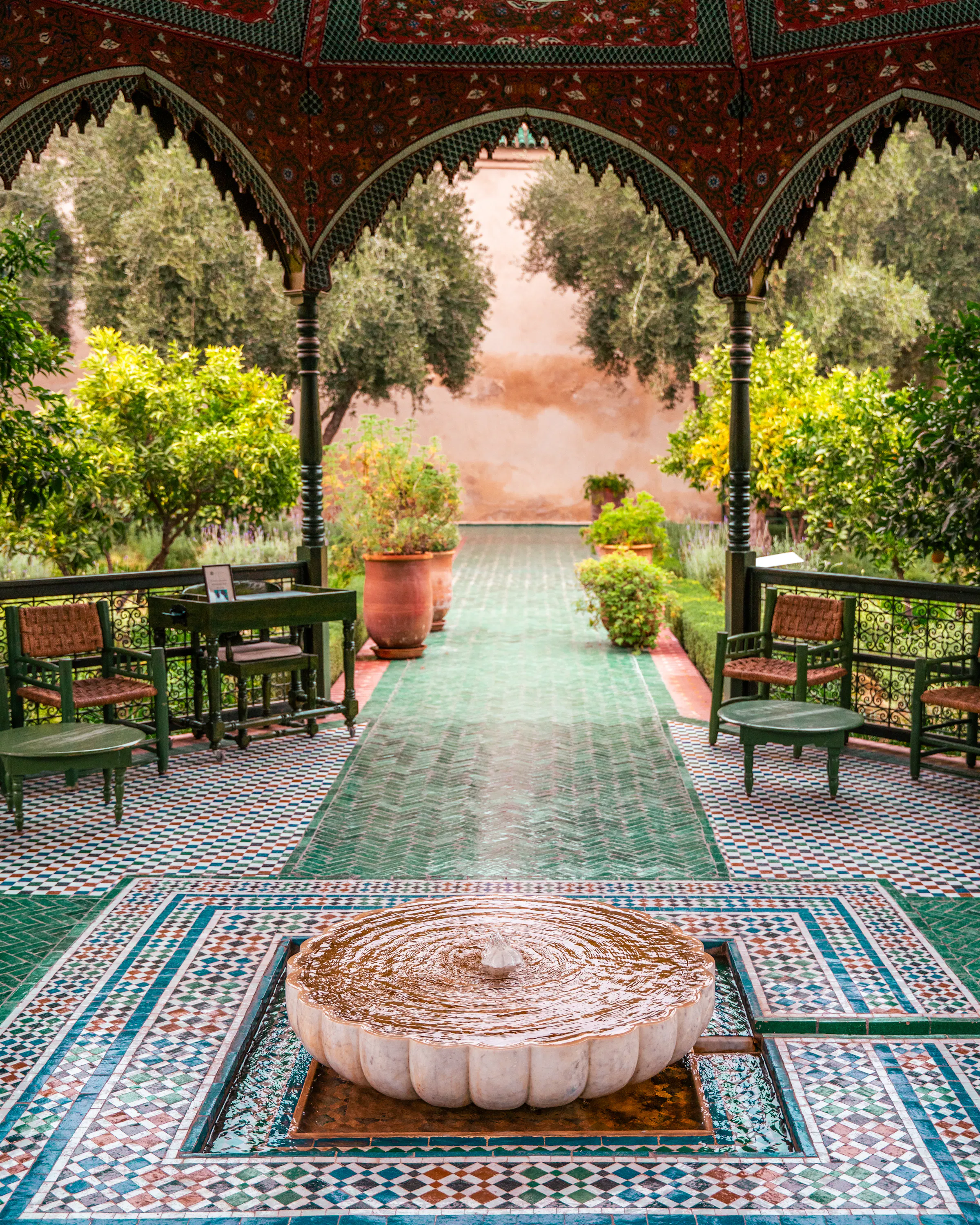 A tiled fountain ripples beneath a patterned pavilion in Marrakech, bordered by green chairs and a formal garden.