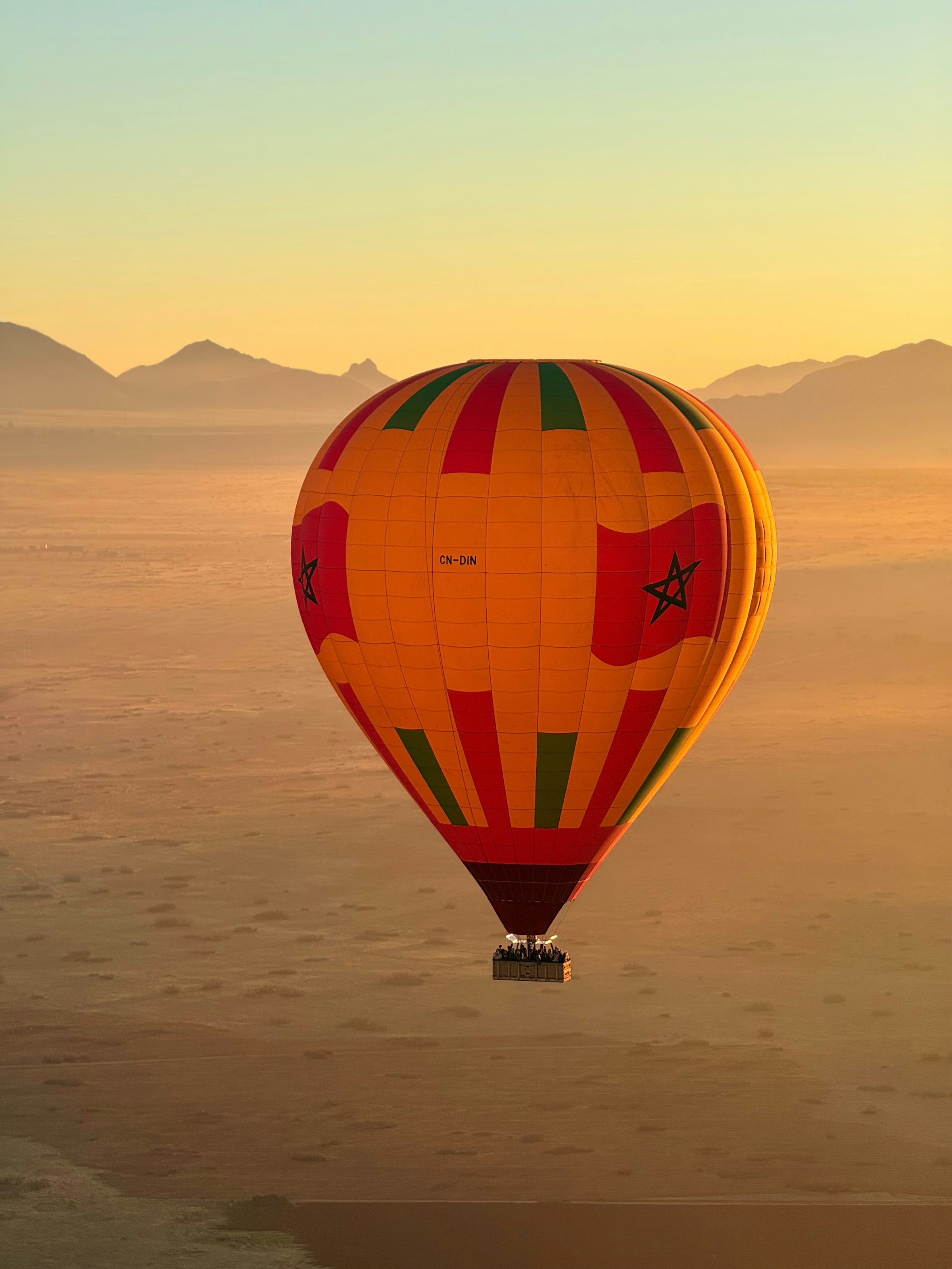 A colorful hot air balloon floats above the desert at sunrise, with soft sand, hazy mountains, and still air below.