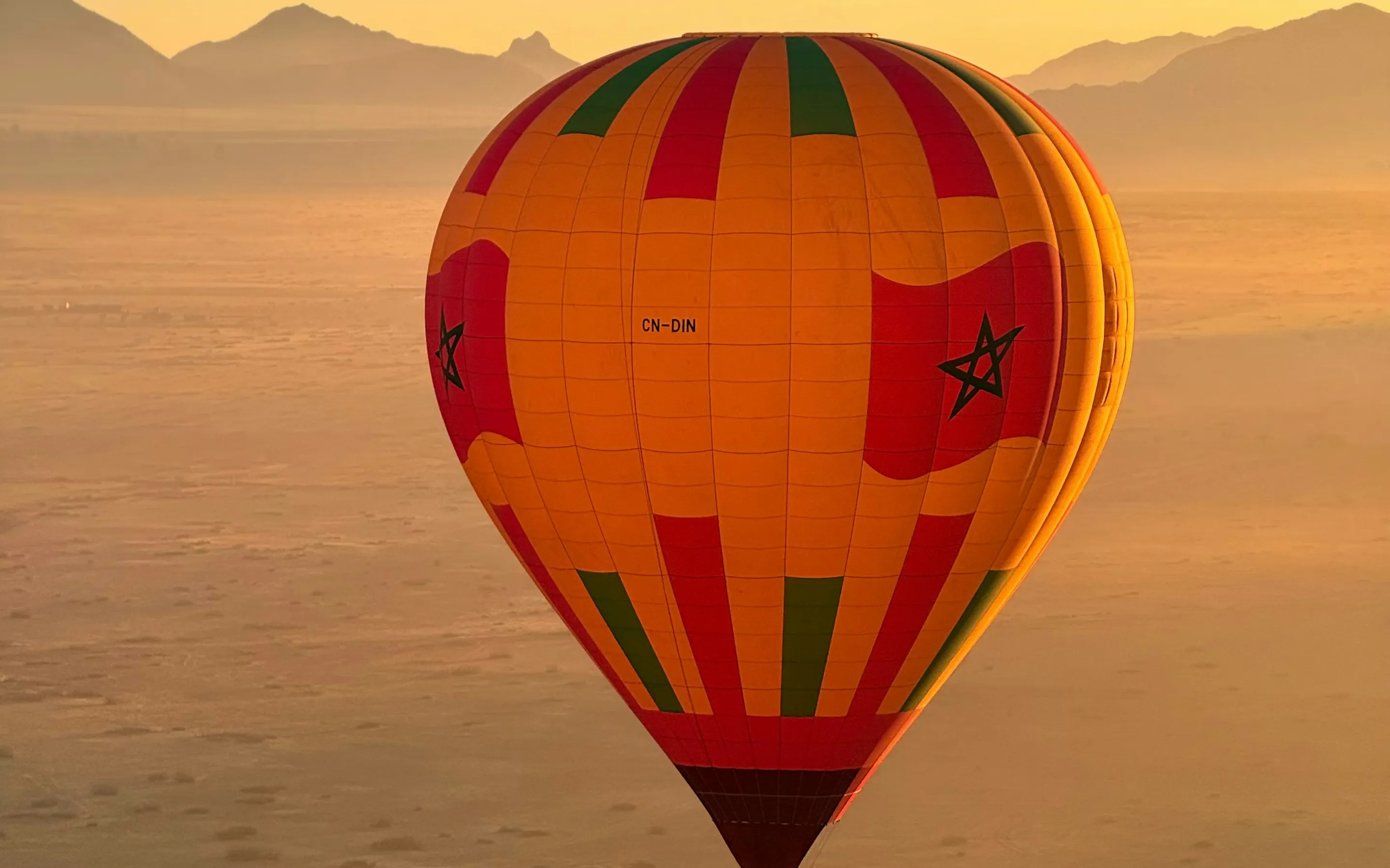 A colorful hot air balloon floats above the desert at sunrise, with soft sand, hazy mountains, and still air below.