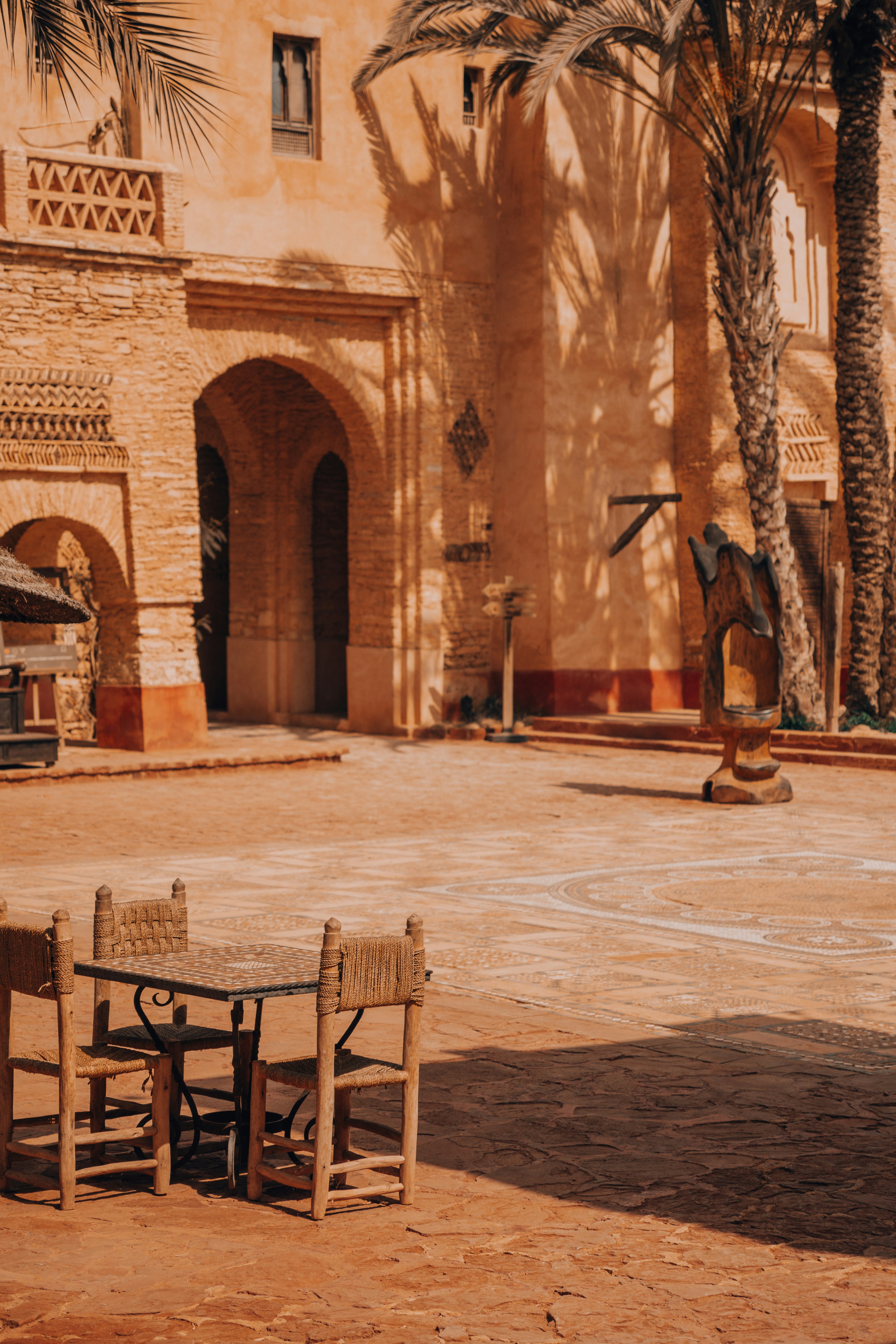 A quiet courtyard sits beneath tall palms, with arched doorways, terracotta walls, and a table set in warm light.