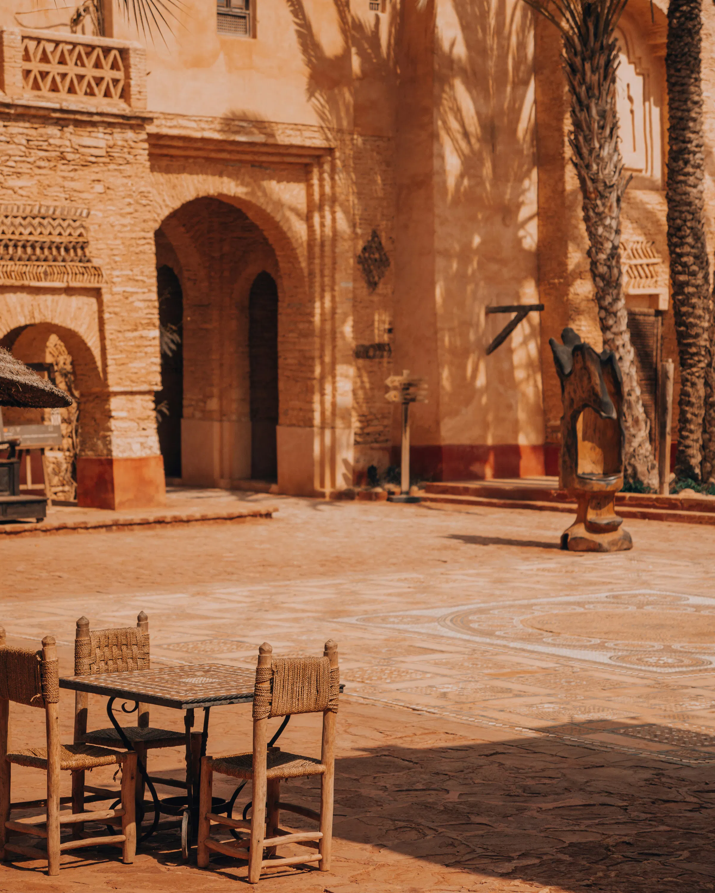 A quiet courtyard sits beneath tall palms, with arched doorways, terracotta walls, and a table set in warm light.