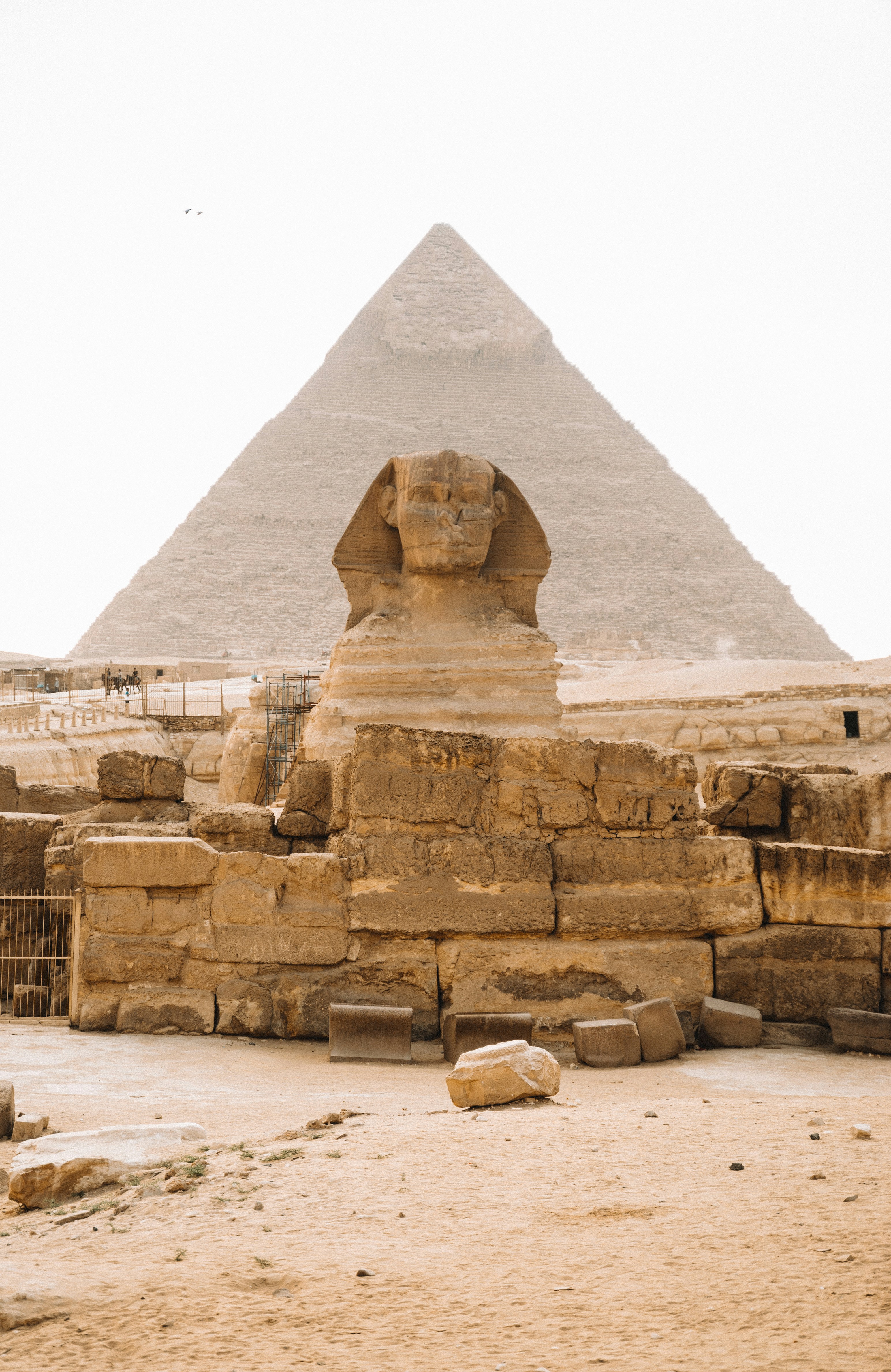 The Great Sphinx stands before a pyramid at Giza, with pale stone blocks, sandy ground, and a hazy white sky.