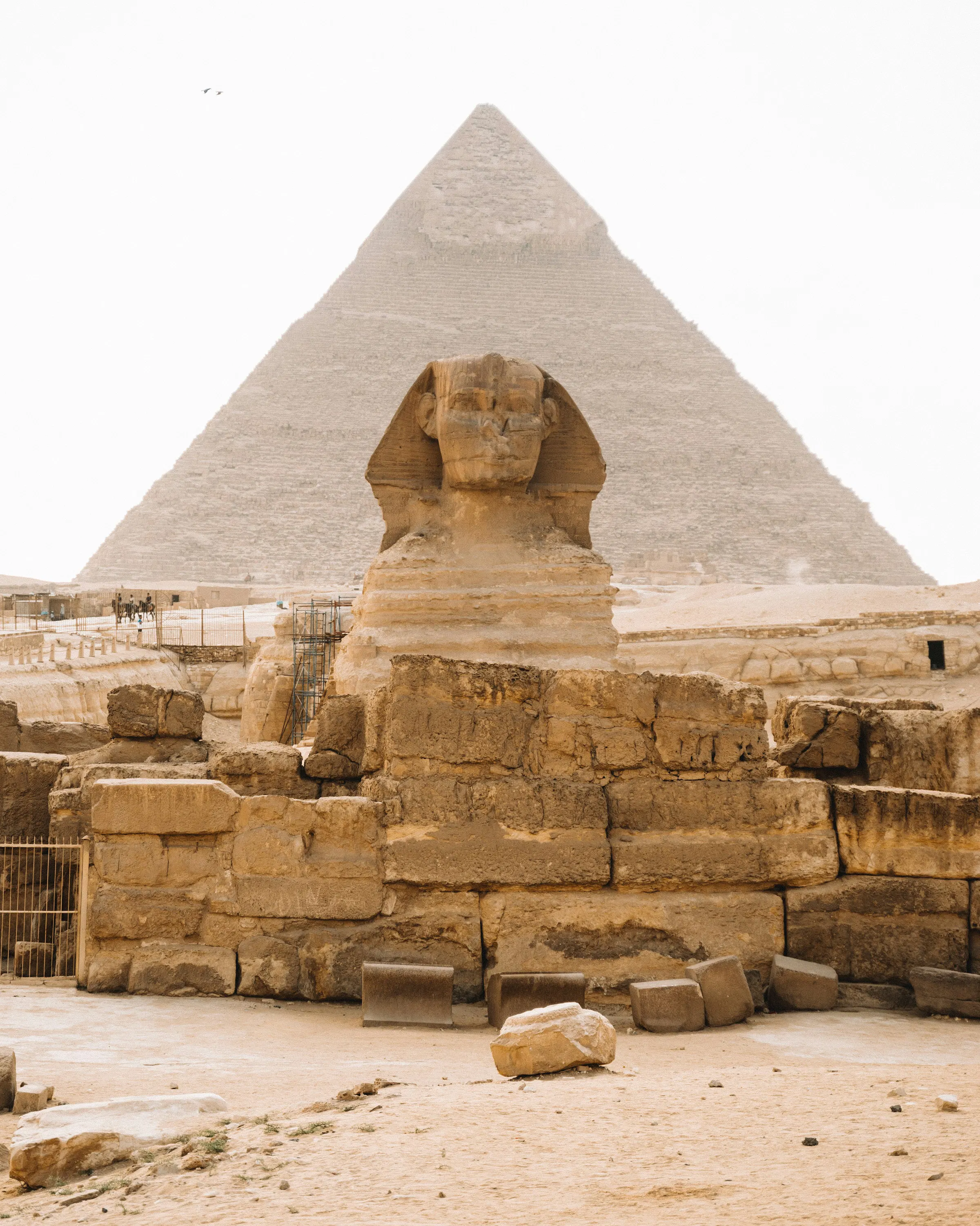 The Great Sphinx stands before a pyramid at Giza, with pale stone blocks, sandy ground, and a hazy white sky.