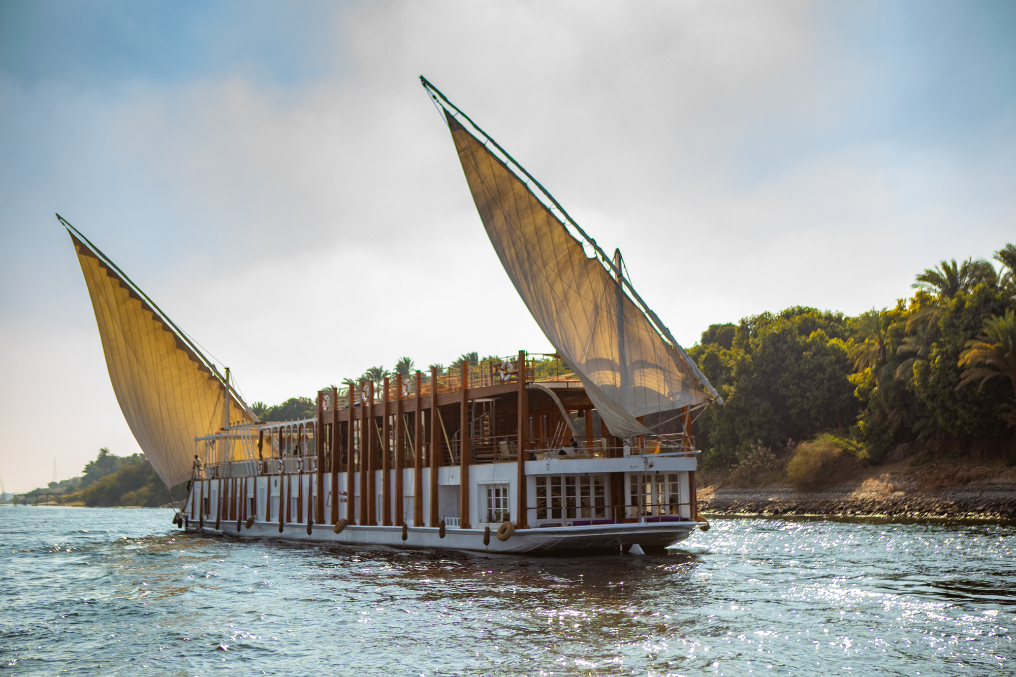 A white dahabiya sails along the Nile beside palm-lined banks, with tall canvas sails and bright water in view.