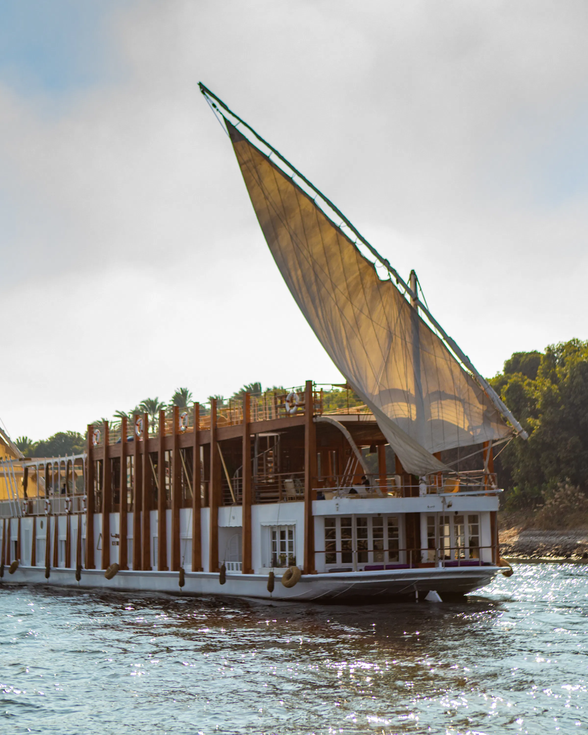 A white dahabiya sails along the Nile beside palm-lined banks, with tall canvas sails and bright water in view.