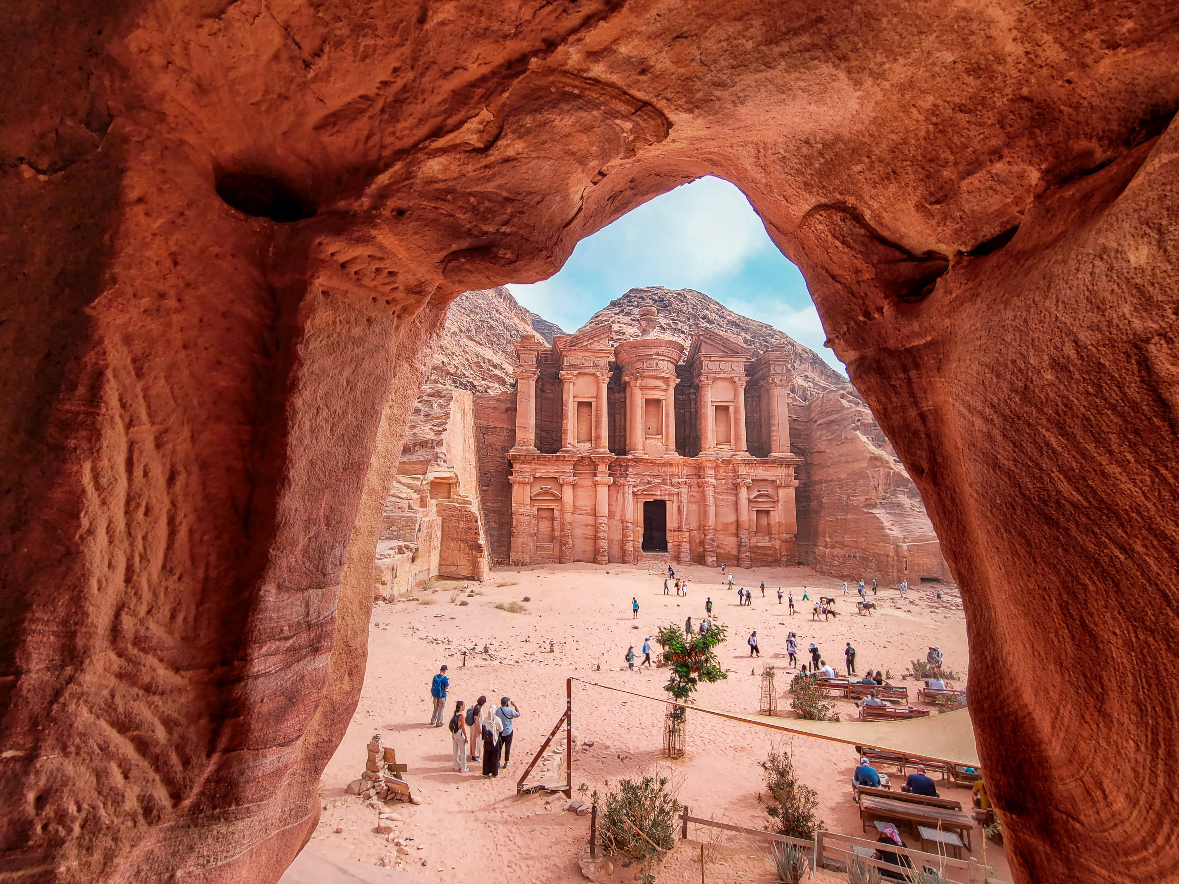 Petra's Monastery rises beyond a red sandstone opening, with visitors scattered across the sandy forecourt below.