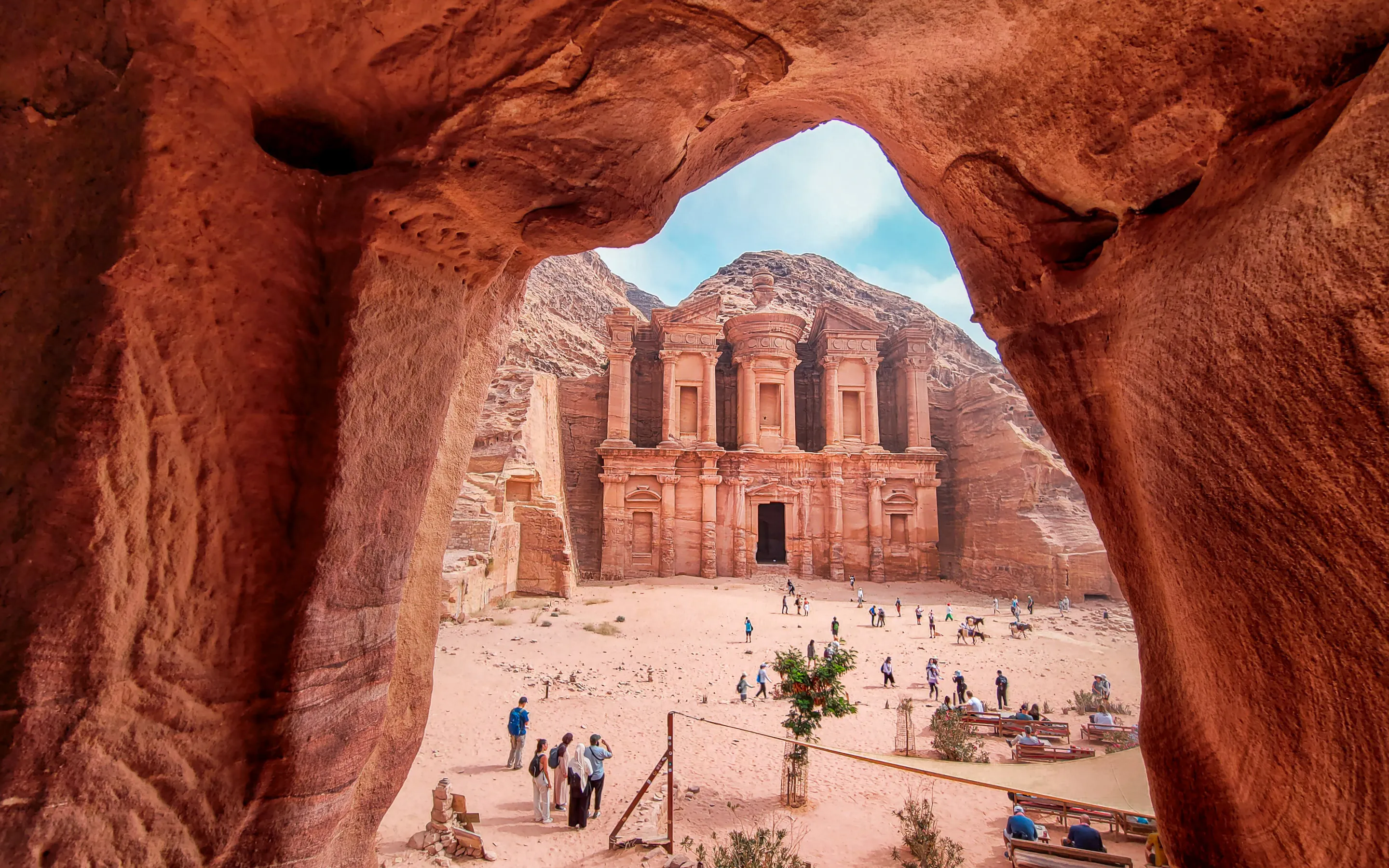 Petra's Monastery rises beyond a red sandstone opening, with visitors scattered across the sandy forecourt below.