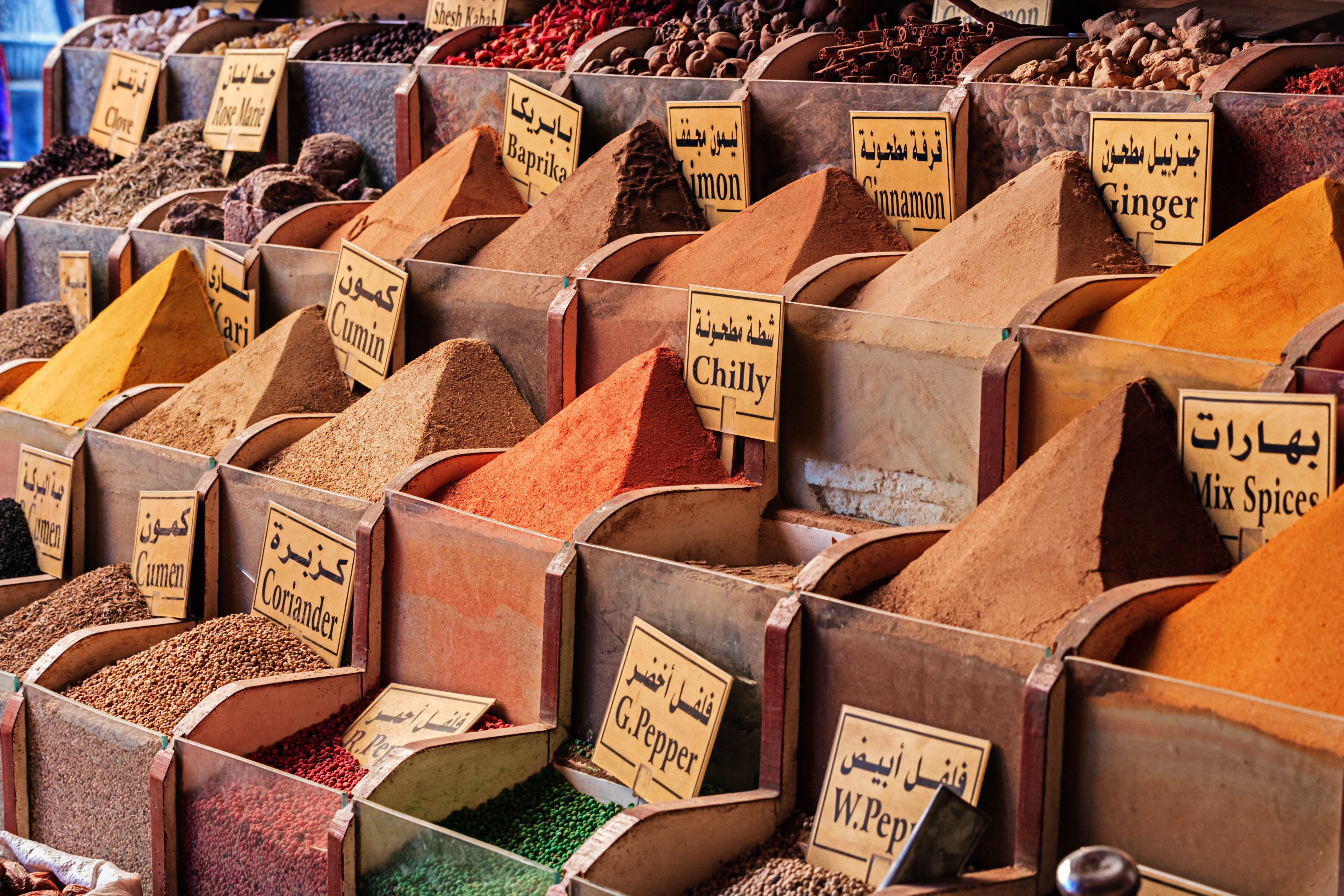 Rows of spices and dried herbs rise in neat pyramids, each labeled in Arabic and English across a market stall.