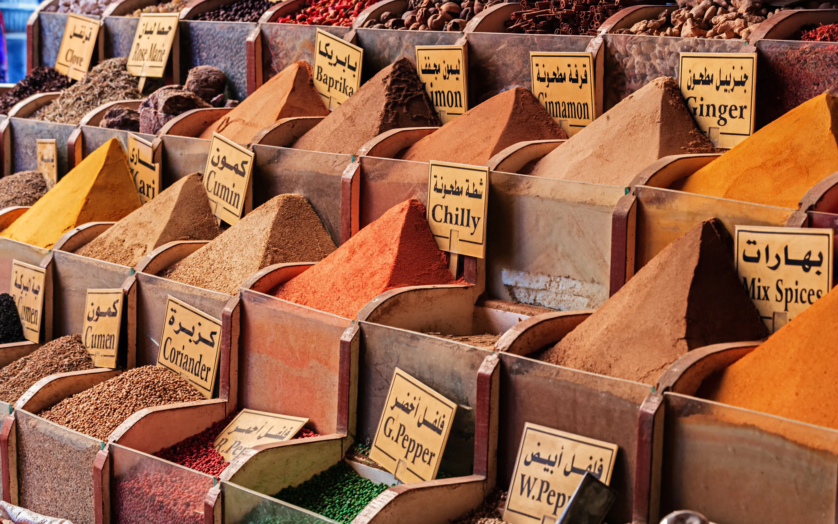 Rows of spices and dried herbs rise in neat pyramids, each labeled in Arabic and English across a market stall.