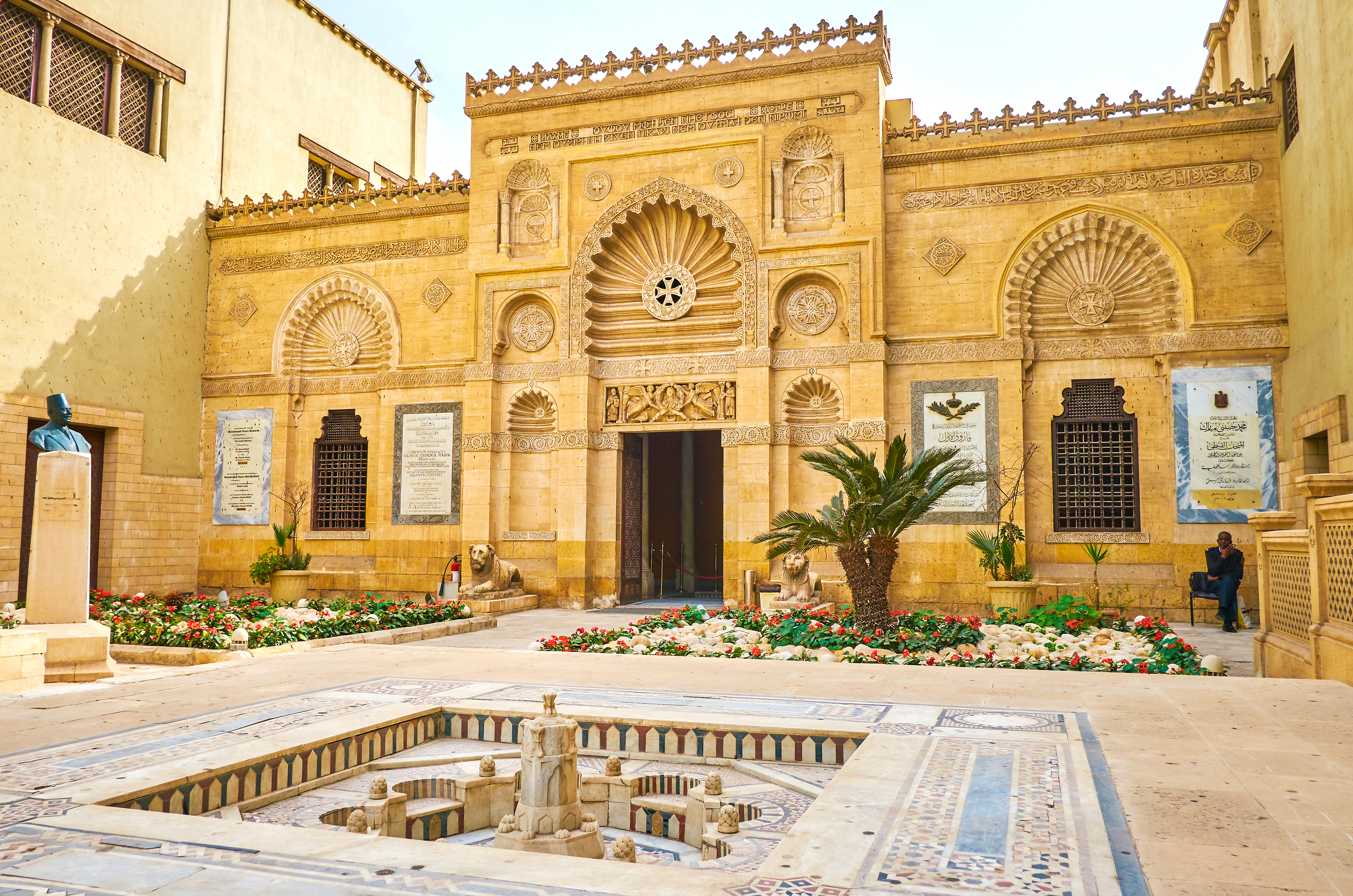 The ornate façade of Cairo's Coptic Museum rises above a tiled courtyard fountain, framed by carved stone details.