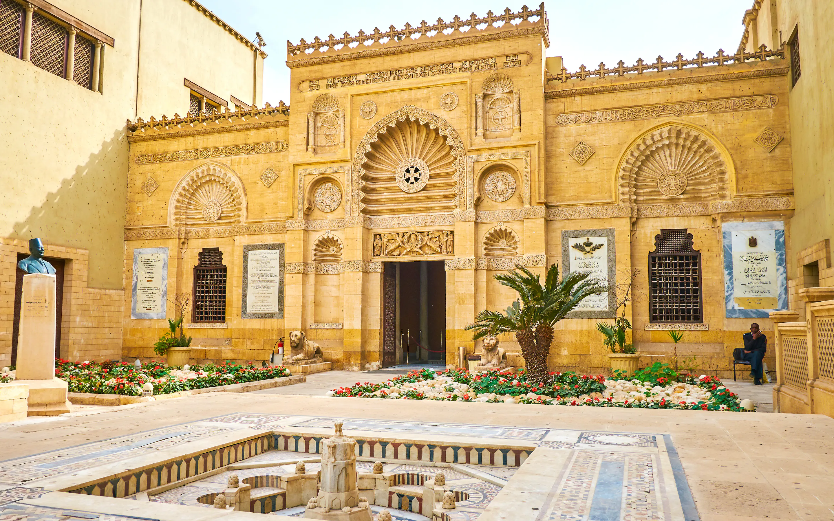 The ornate façade of Cairo's Coptic Museum rises above a tiled courtyard fountain, framed by carved stone details.