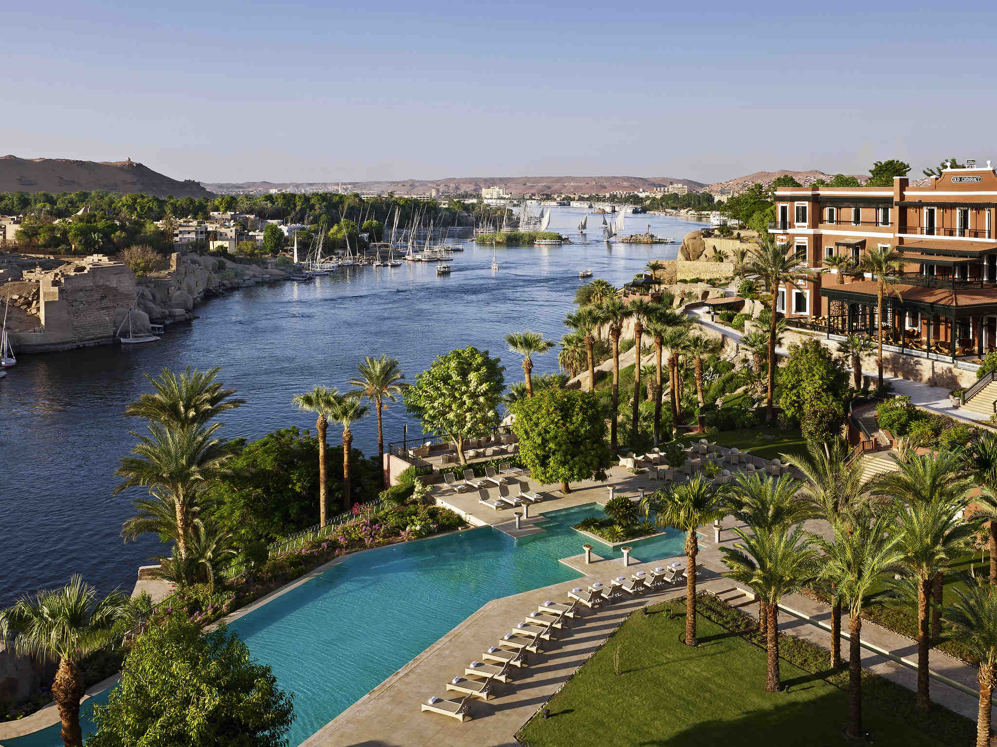 The pool and gardens of the Old Cataract overlook the Nile in Aswan, with sailboats drifting beyond.