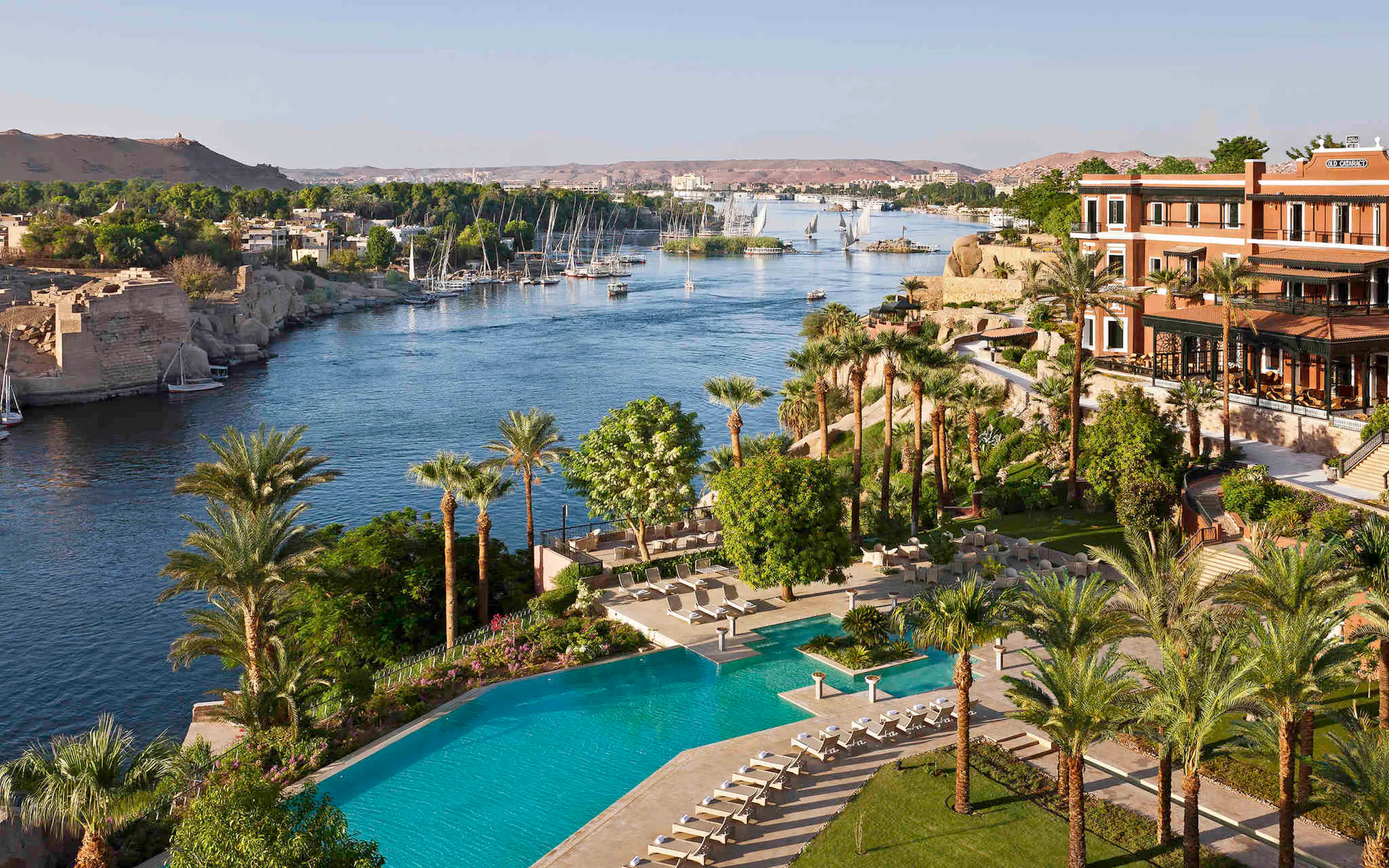 The pool and gardens of the Old Cataract overlook the Nile in Aswan, with sailboats drifting beyond.