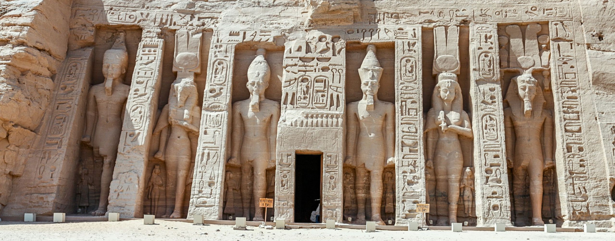 Colossal carved figures and hieroglyphs line the stone façade of Abu Simbel, rising above a bright sandy forecourt.