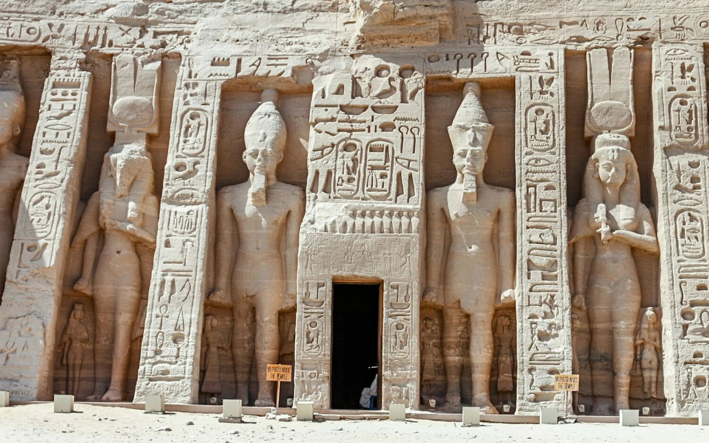 Colossal carved figures and hieroglyphs line the stone façade of Abu Simbel, rising above a bright sandy forecourt.
