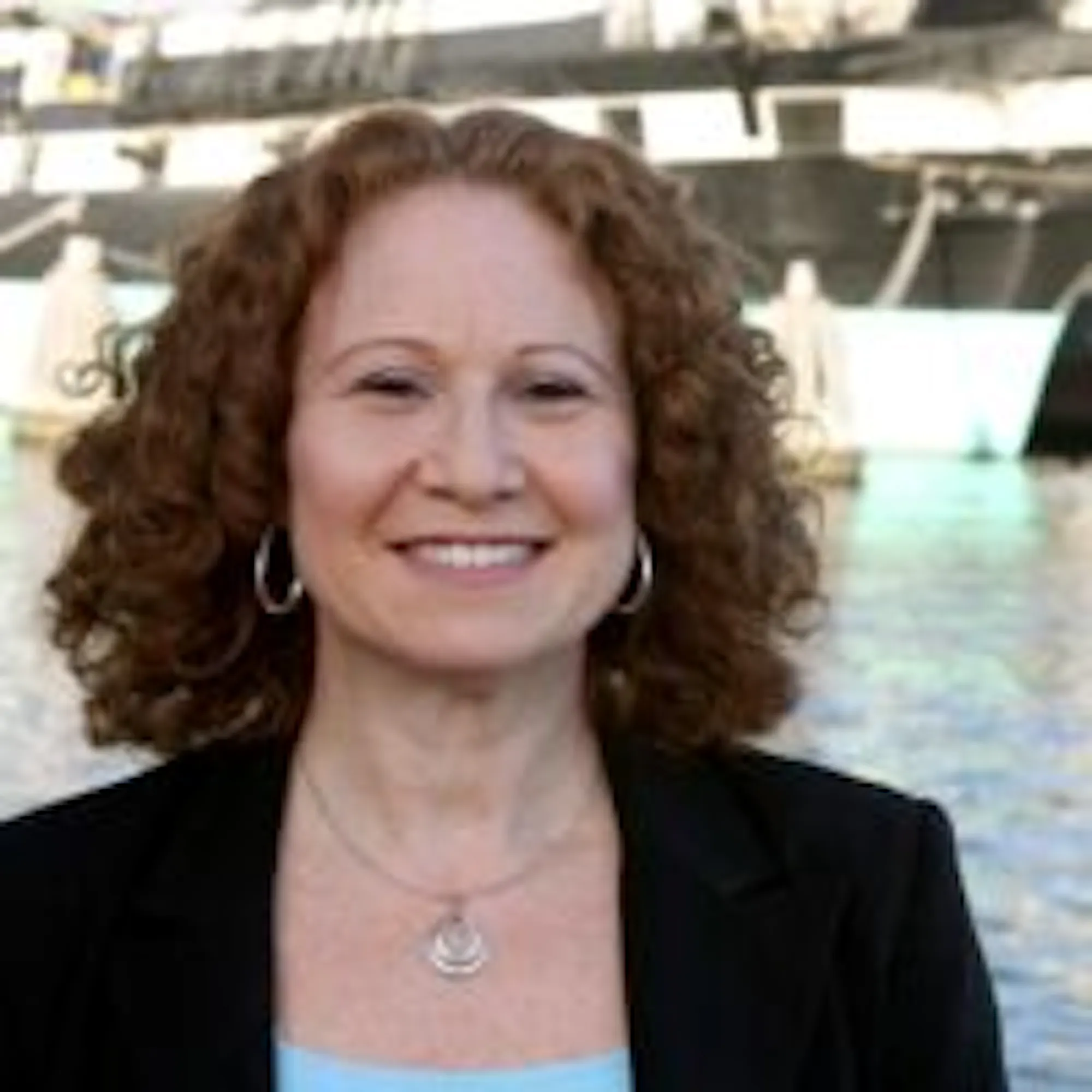 A woman with curly red hair wearing a black blazer and blue top stands in front of a marina and smiles at the camera. 