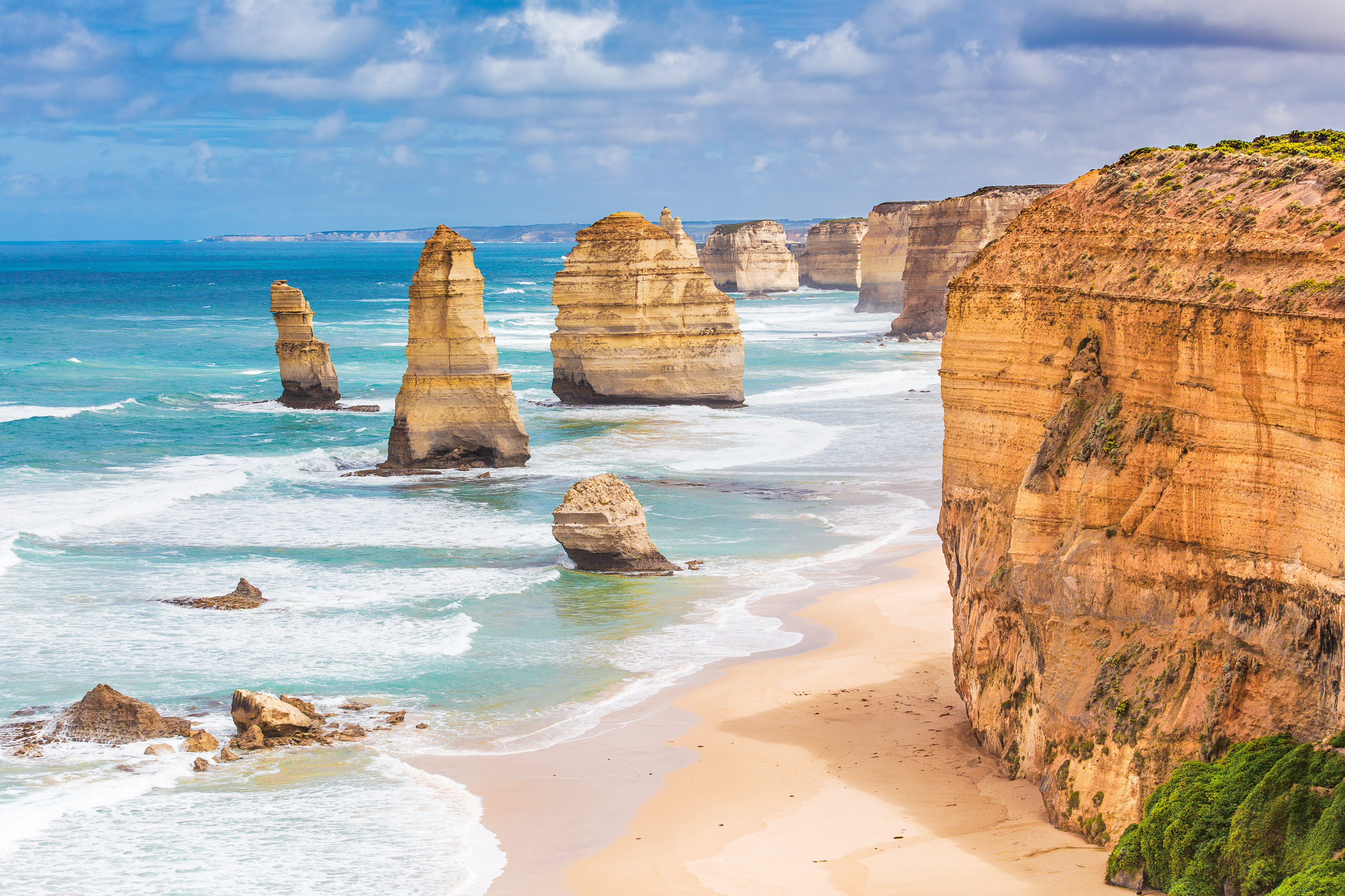 Golden sea stacks rise beside the Twelve Apostles as turquoise waves roll toward a long sandy beach below.