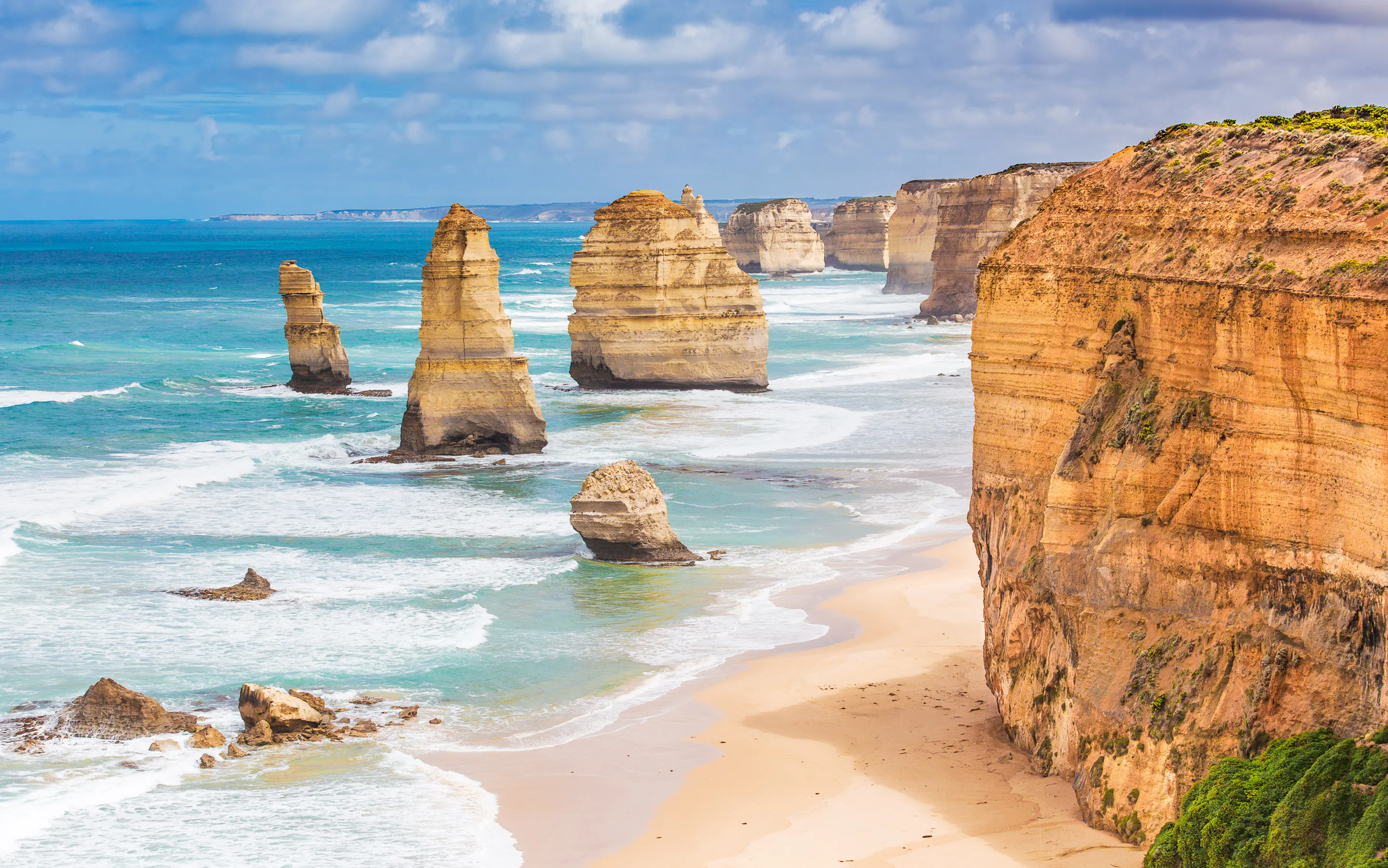 Golden sea stacks rise beside the Twelve Apostles as turquoise waves roll toward a long sandy beach below.