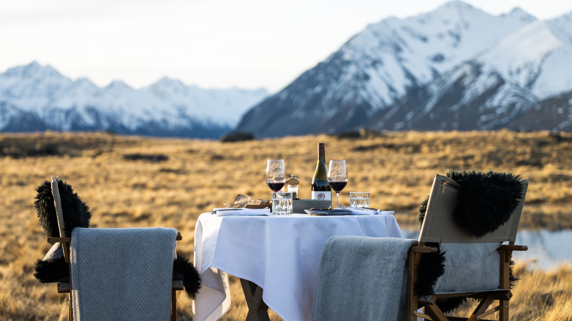 A linen-covered table for two sits beside a still lake, with snowy peaks and golden grass stretching into the distance.
