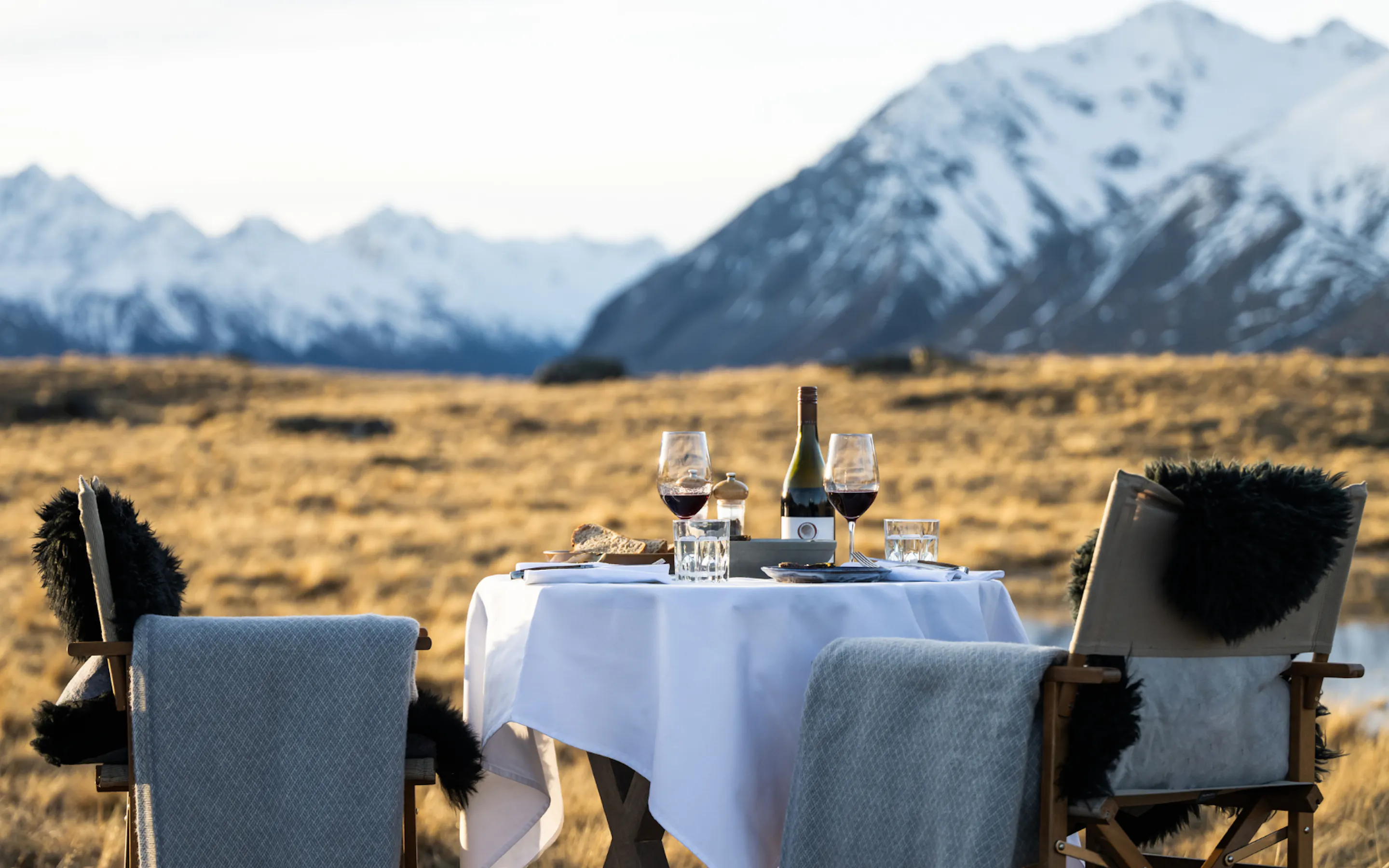 A linen-covered table for two sits beside a still lake, with snowy peaks and golden grass stretching into the distance.