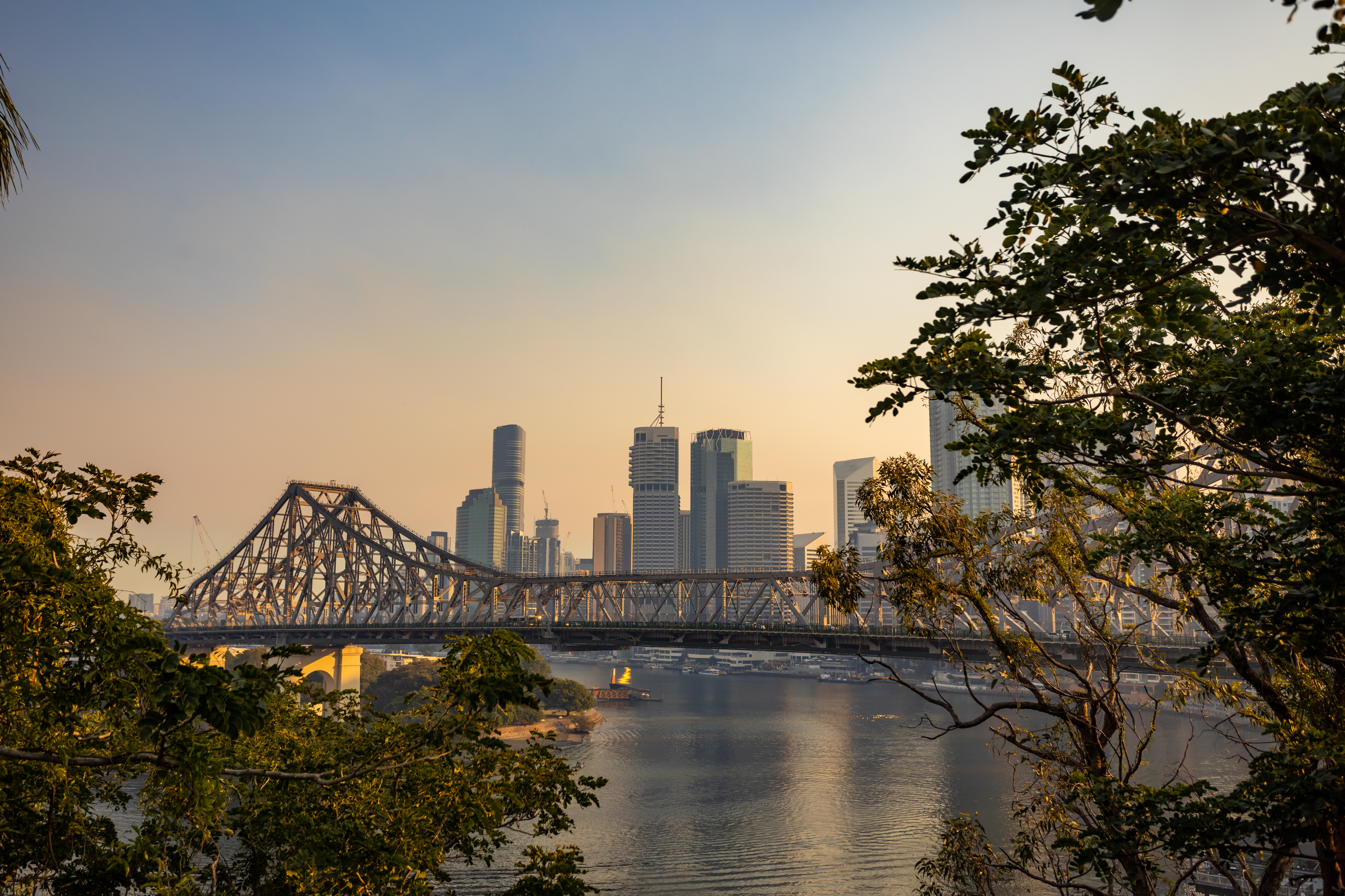 The Story Bridge spans the Brisbane River at sunset, with the city skyline glowing behind riverside trees.