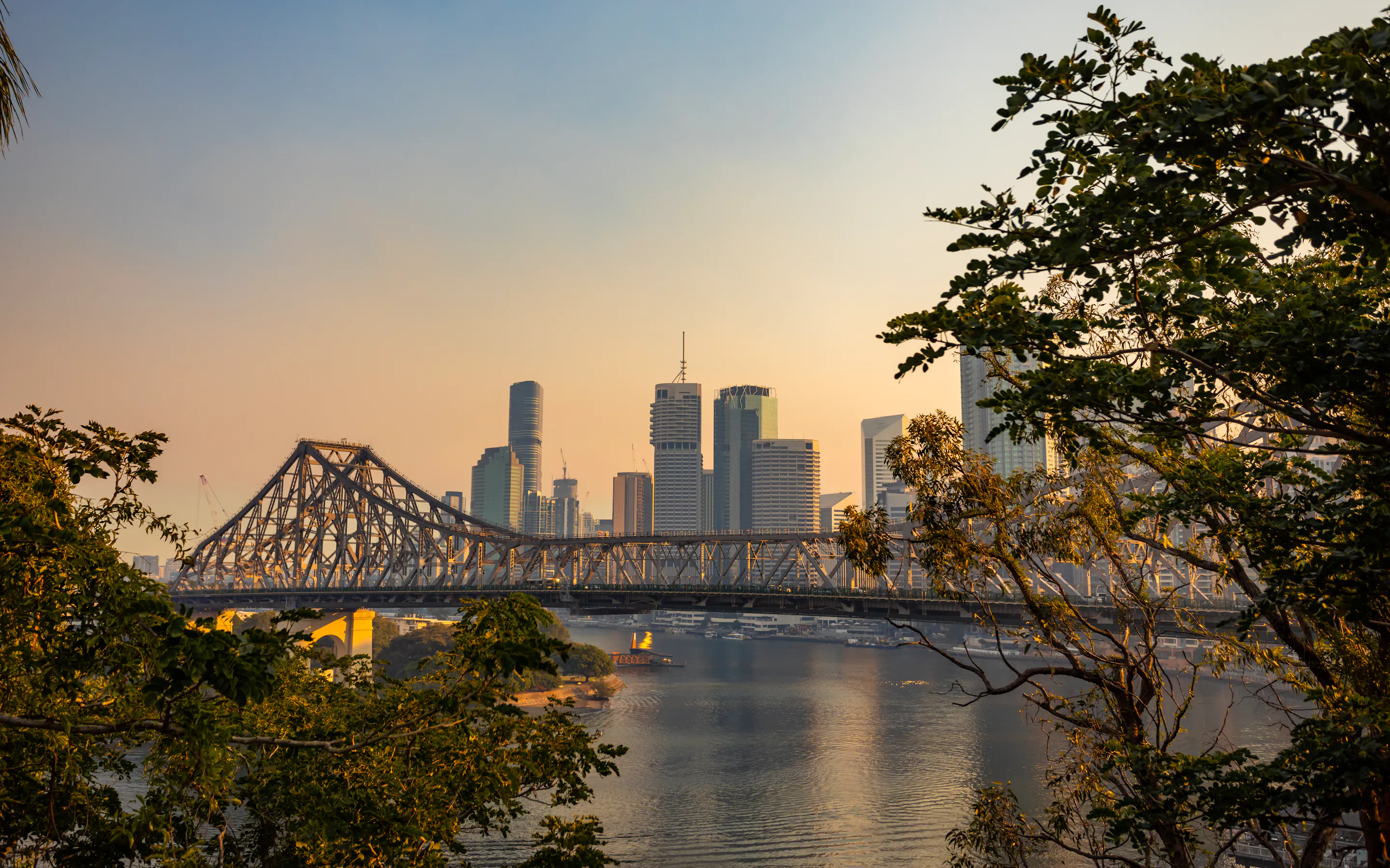 The Story Bridge spans the Brisbane River at sunset, with the city skyline glowing behind riverside trees.