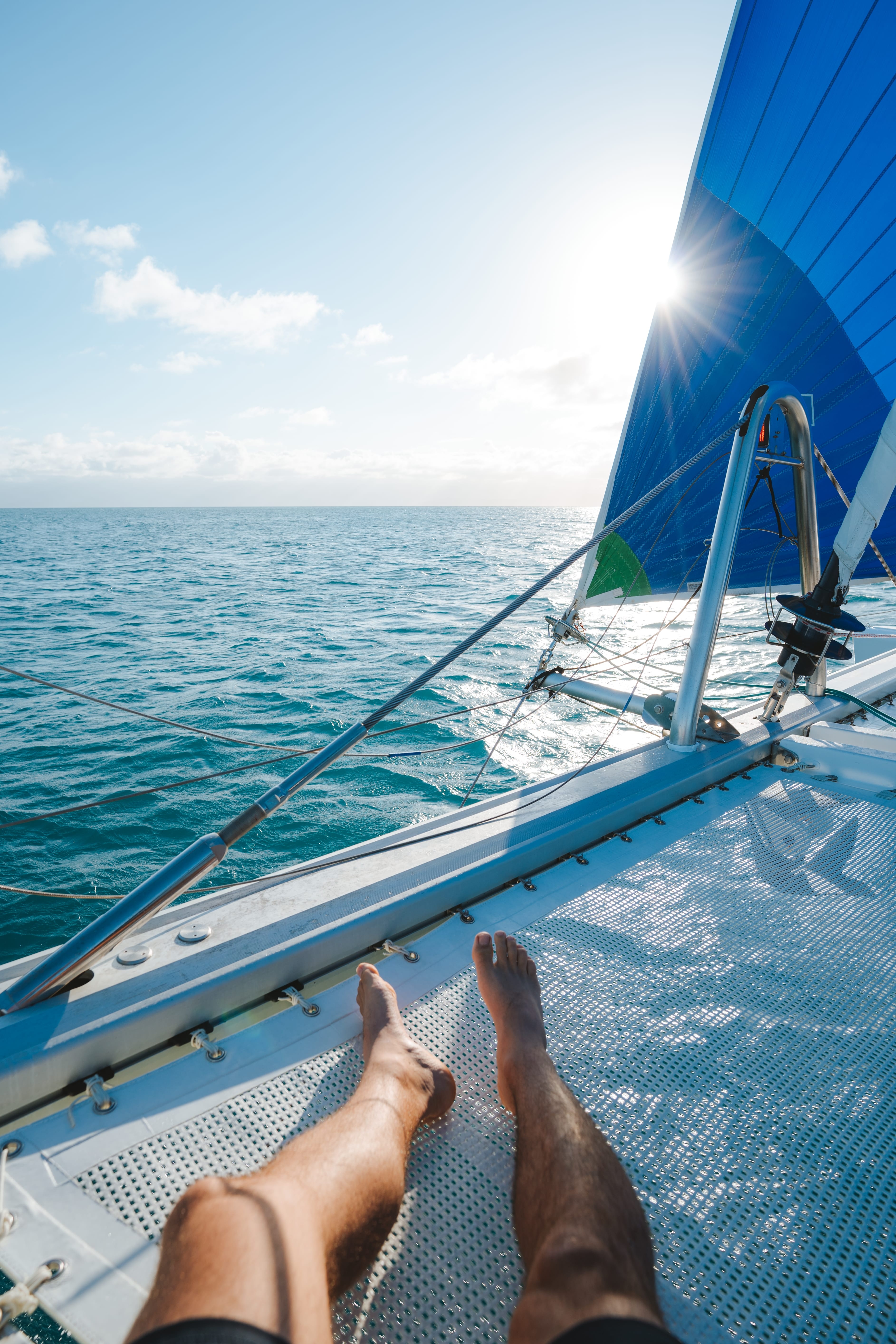 Bare feet rest on a catamaran net above clear water, with a blue sail near Low Isles.