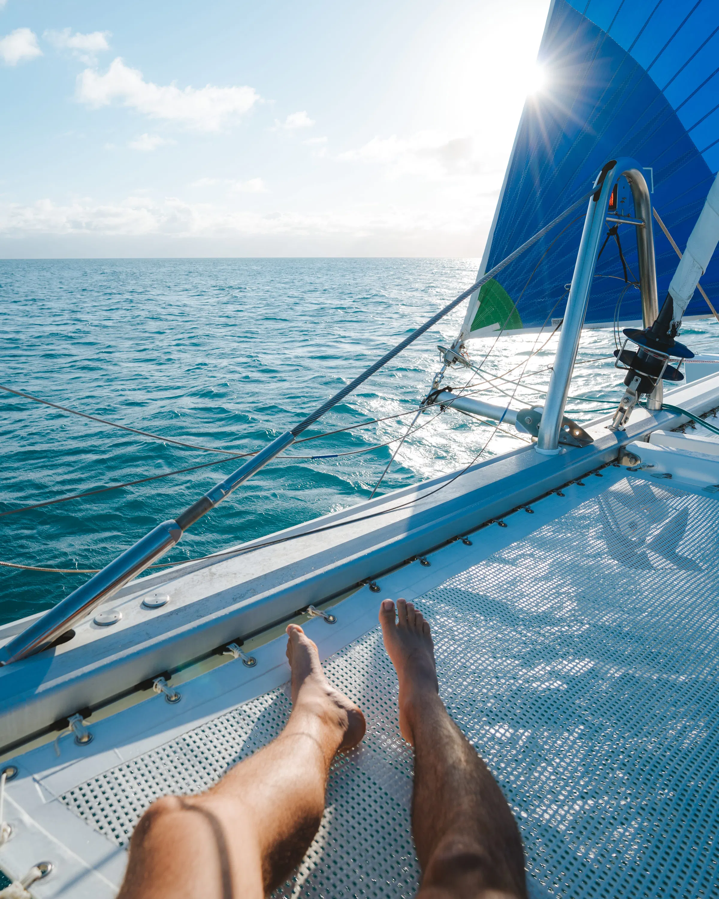 Bare feet rest on a catamaran net above clear water, with a blue sail near Low Isles.